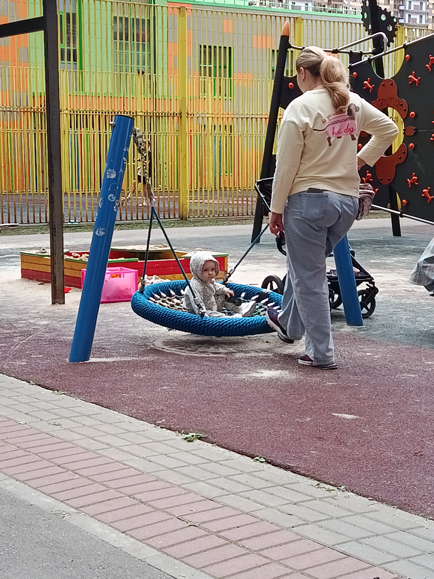 A woman stands behind a baby in a hanging swing at a playground. The woman is wearing a light-colored long-sleeved shirt with a graphic of a dachshund on the back and blue jeans. The baby is wearing a gray hooded outfit and is seated in a blue rope swing. Behind them is a colorful fence and playground equipment. In the foreground, a red and gray patterned surface covers the ground.