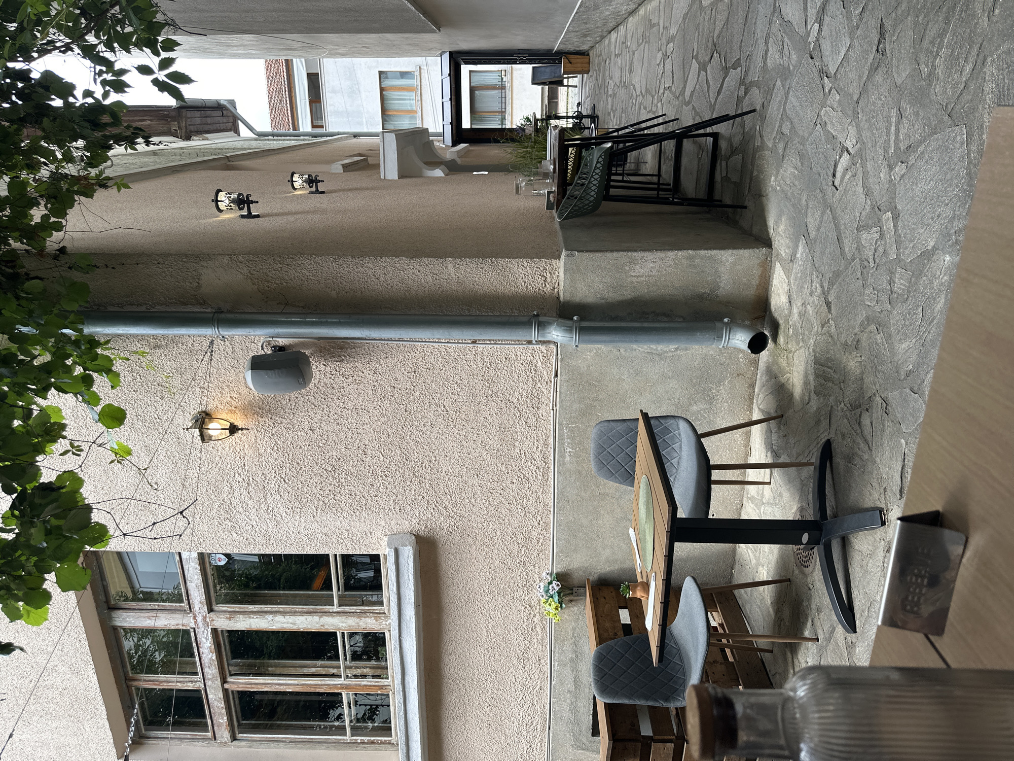 This image depicts the outdoor seating area of a cafe or restaurant located in the city of Sofia, Bulgaria. The scene is set in a narrow alleyway between buildings. In the foreground, a glass bottle with a cork stopper rests on a wooden surface. A metal "RESERVE" sign is positioned in front of this surface.
Further into the alley, a small square wooden table is set with two grey, diamond-patterned chairs. The table has a green placemat, cutlery, and a small potted plant. Behind this table, a wooden structure is visible. On the wall of the building to the left, a window with multiple panes and weathered wooden frames is present. A wall-mounted light fixture illuminates the textured, pale pink exterior wall, and a drainpipe runs vertically down the corner of the building.
Further down the alley, a high table is accompanied by three black bar stools. More potted plants and a glass jar filled with water and reeds are on this table. The alley floor is paved with irregular grey flagstones. In the background, more buildings and doorways are visible, including an entrance with a dark awning. Green foliage hangs from above, partially obscuring the top of the scene. The overall atmosphere suggests a quiet, possibly overcast day.