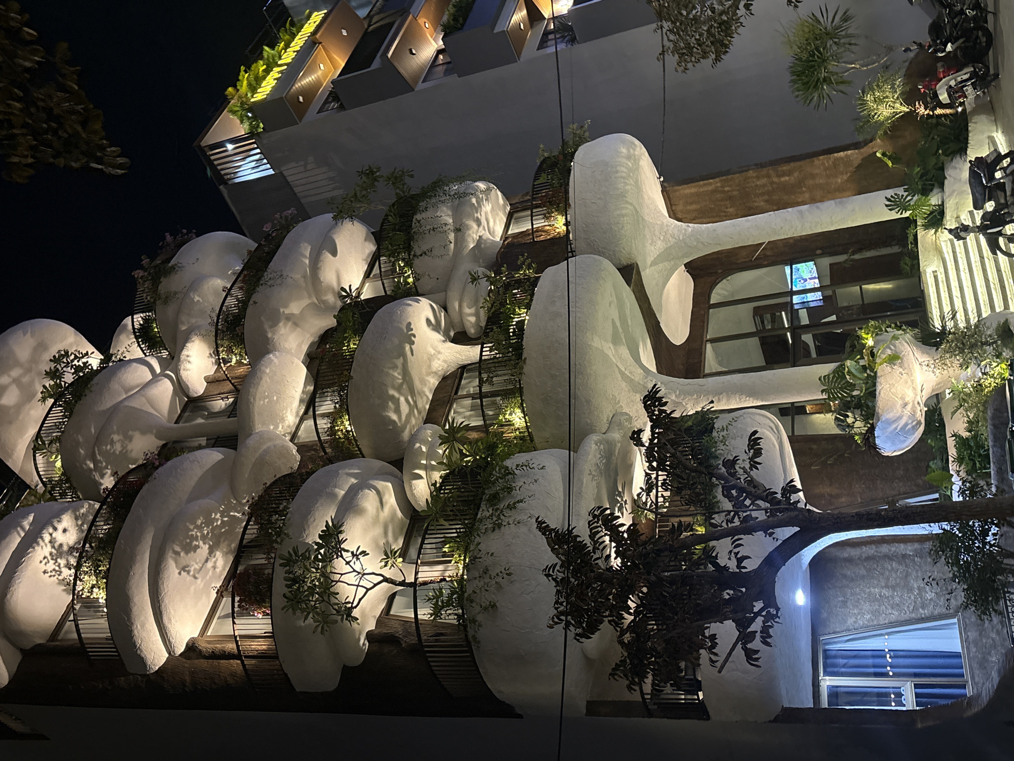This media file depicts the exterior of a uniquely designed building at night in Da Nang, Vietnam. The building features an avant-garde architectural style with large, bulbous, white structures that resemble natural forms or abstract sculptures. These elements are integrated with balconies and windows, suggesting it is a multi-story residential or commercial property. Lush greenery and plants are strategically placed on and around these structures, creating a biophilic design that softens the sharp architectural lines.
The scene is illuminated by artificial lighting, highlighting the building's unusual contours and the plants. The time of day is clearly night, indicated by the dark sky. No specific weather conditions are apparent, but the overall ambiance suggests a calm evening. The location is identified as Da Nang, Vietnam, though no specific landmarks are visible.
The primary subjects are the building itself and its distinctive design. There are no visible people or any clear indication of an event taking place. The notable details are the organic, sculptural forms of the architecture, the integration of plant life, and the nighttime illumination. The design evokes a sense of wonder and perhaps artistic expression, with no discernible emotions expressed by the scene itself. There is visible text on the upper floors of the building, appearing to be a yellow illuminated sign that reads "MƯỜNG THANH".