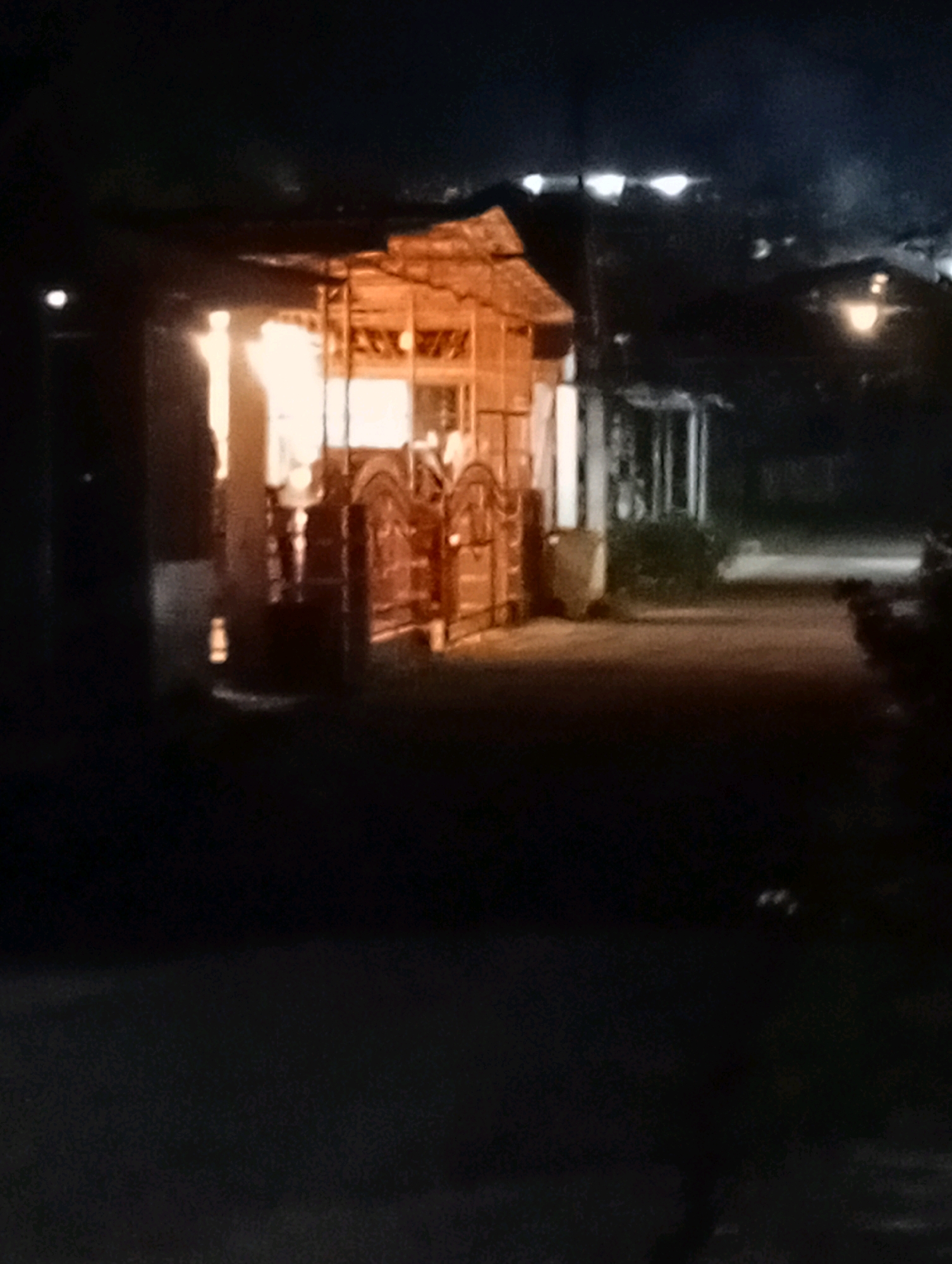 The image depicts a dark outdoor scene at night, likely in a residential or commercial area of the City of Gunungsitoli, Indonesia. The focal point is a small, illuminated structure with a thatched roof and wooden walls, possibly a kiosk or a small shop. Bright interior lighting casts a warm glow from within. In front of this structure, there are two large, ornate wooden doors, possibly part of the entrance or decorative elements. The ground in front appears to be a paved surface, possibly a pathway or road, which is dimly lit. To the right of the structure, there is a visible pathway or road with some vegetation and street lighting in the background. The overall atmosphere is quiet and somewhat mysterious due to the low light conditions.