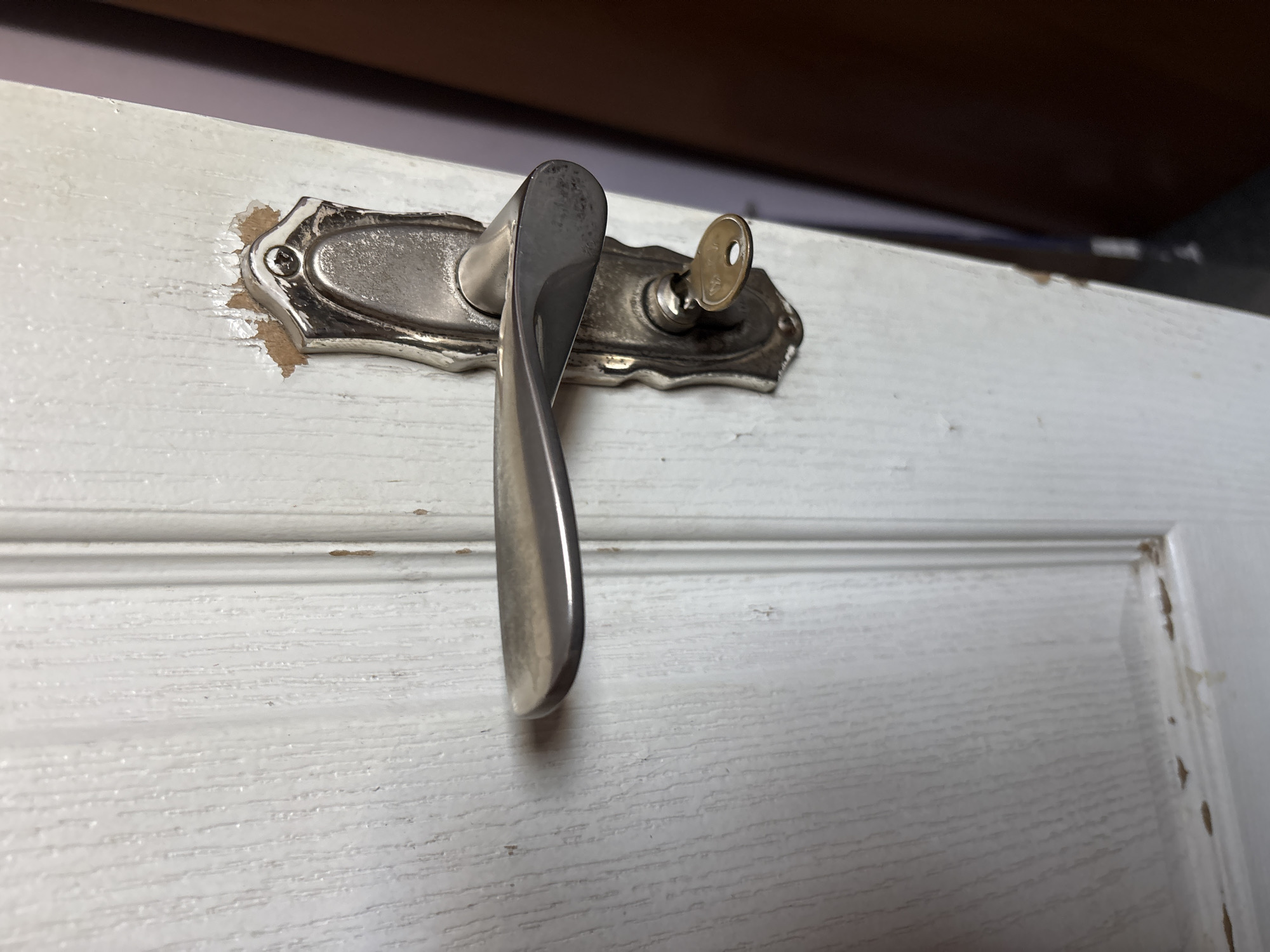 The image is a close-up, eye-level, medium shot of a white, wooden door with a silver-colored doorknob and a key inserted into the lock. The door appears to be old and shows signs of wear, with chipped paint around the doorknob. The wood grain is visible through the white paint, suggesting a textured surface. A dark brown door or wall is visible in the background to the right, out of focus. The lighting is moderate, casting subtle shadows. There are no people or animals present in the frame. The scene is static.