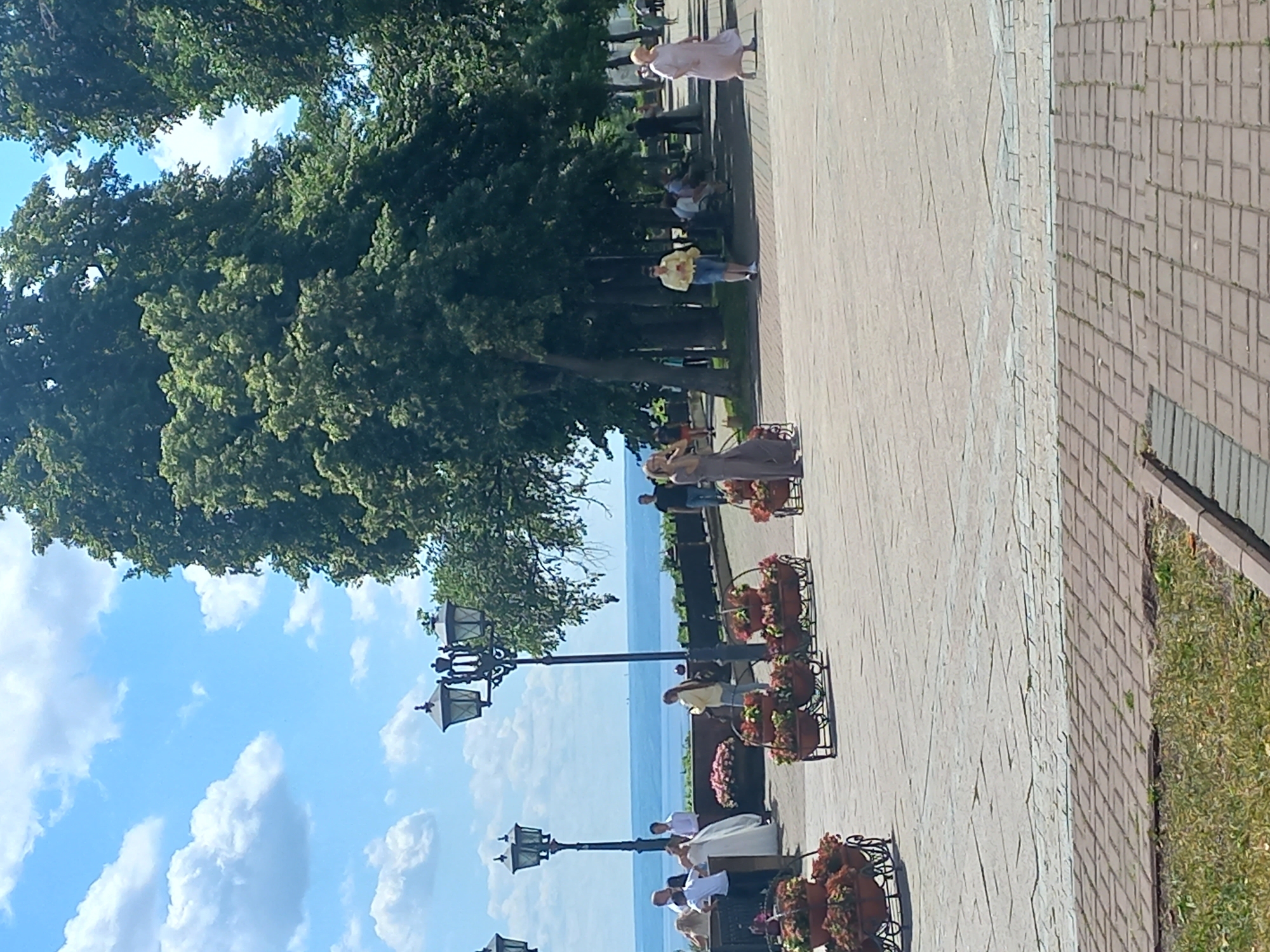 The image depicts a sunny day at a waterfront promenade in Ulyanovsk, Russia. In the foreground, a paved walkway made of light grey rectangular stones transitions to a darker brick border, with a patch of dry grass to the left. Flower beds filled with red and pink blooms are strategically placed along the walkway, some on wheeled carts.
Further back, several people are visible. A group appears to be gathered near a wedding dress, suggesting a celebration. Among them, a woman in a long, flowing grey dress stands apart, looking towards the water. Another woman in a yellow shirt and blue shorts walks across the promenade. Further down, a woman in a light pink dress is walking, and other individuals are seated on benches or strolling.
The background features a wide expanse of water, likely the Volga River, under a blue sky dotted with white clouds. Tall, mature trees with lush green foliage line the promenade, providing shade. Several ornate black lampposts are also present, typical of the city's public spaces. The overall scene is one of leisure and activity in an urban park setting.
