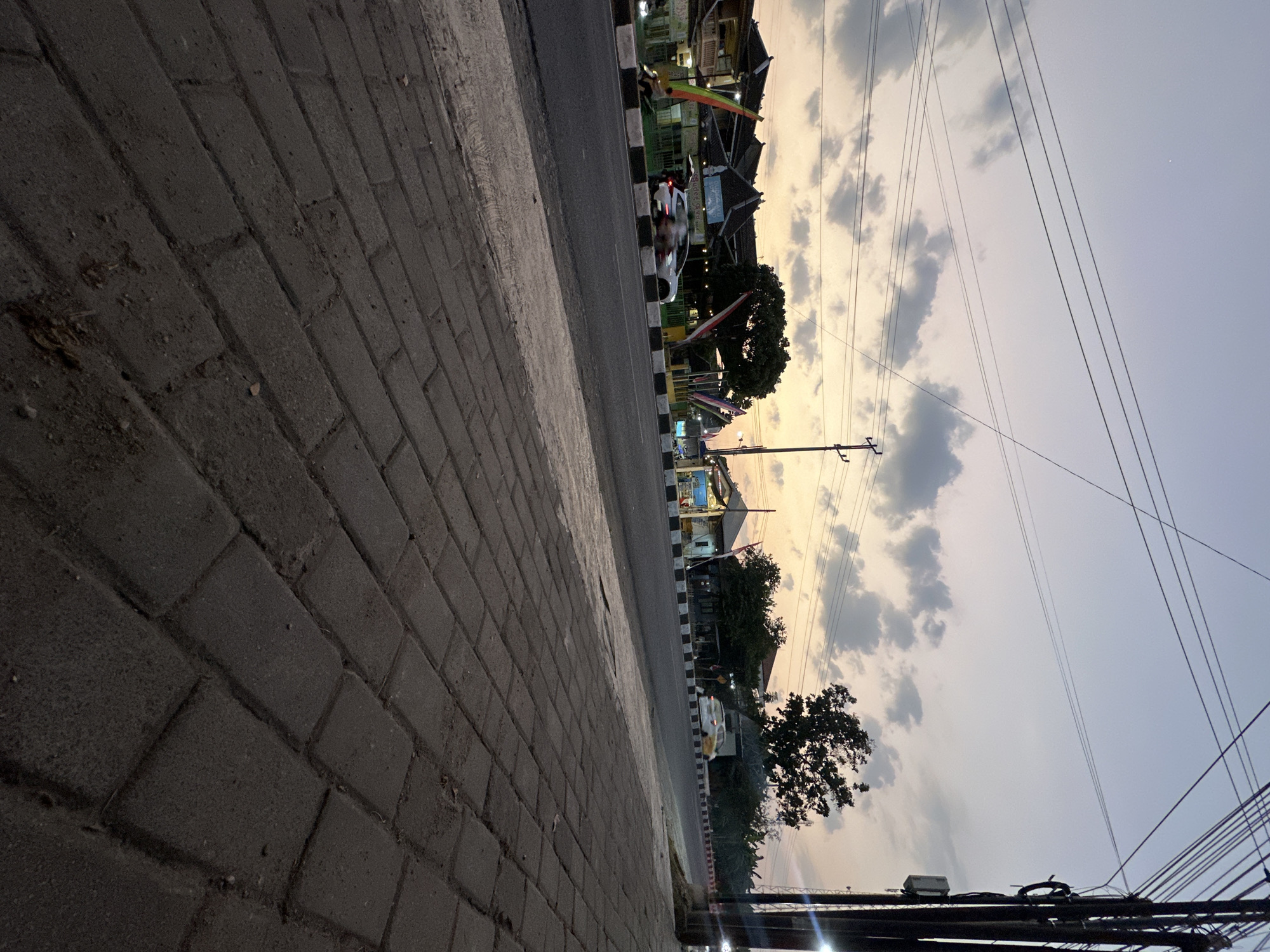 The image depicts a street scene in Bulakrejo, Indonesia, at dusk. A paved sidewalk, made of dark grey rectangular bricks, occupies the foreground and extends to the right. The sidewalk is slightly elevated from the road and shows signs of wear, with some dirt and debris scattered on its surface.
The road itself is asphalt, with a white and black striped border. Several vehicles are visible on the road, including a white car in motion in the middle ground, and a motorcycle with a rider on the left side, also in motion. Some of these vehicles are blurred, suggesting movement.
Along the side of the road, there are buildings with traditional Indonesian architecture, featuring dark tiled roofs. Some buildings appear to be shops or food stalls, with awnings and signs. Decorative flags with red and green elements are also displayed in front of some establishments. A few trees are present, one with a dense canopy near the center-right of the frame.
The sky above is a gradient of colors, transitioning from a pale yellow and orange near the horizon to a soft purple and grey higher up. Scattered clouds are visible, catching the fading light. Overhead, multiple electrical wires are strung across the sky, supported by utility poles. One prominent utility pole, with several insulators and cables, stands on the far right side of the image. The overall lighting suggests late evening or early morning.