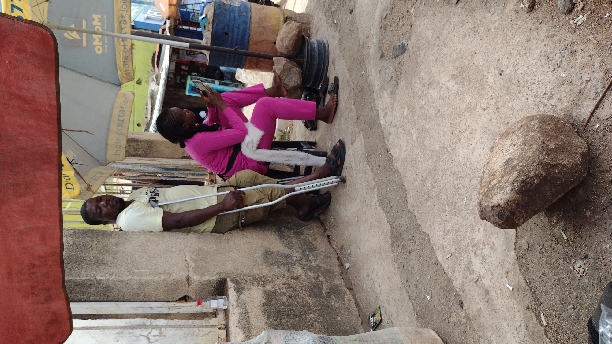 The image depicts a street scene in Jos, Nigeria. A man with a broken leg uses crutches while lying on what seems to be a concrete ledge. A woman, wearing a pink suit and a white scarf, is seated nearby using a smartphone. They are under a large sun umbrella, likely to provide shade from the sun. Debris and litter are present on the ground. A large rock is on the edge of the pavement near the street.