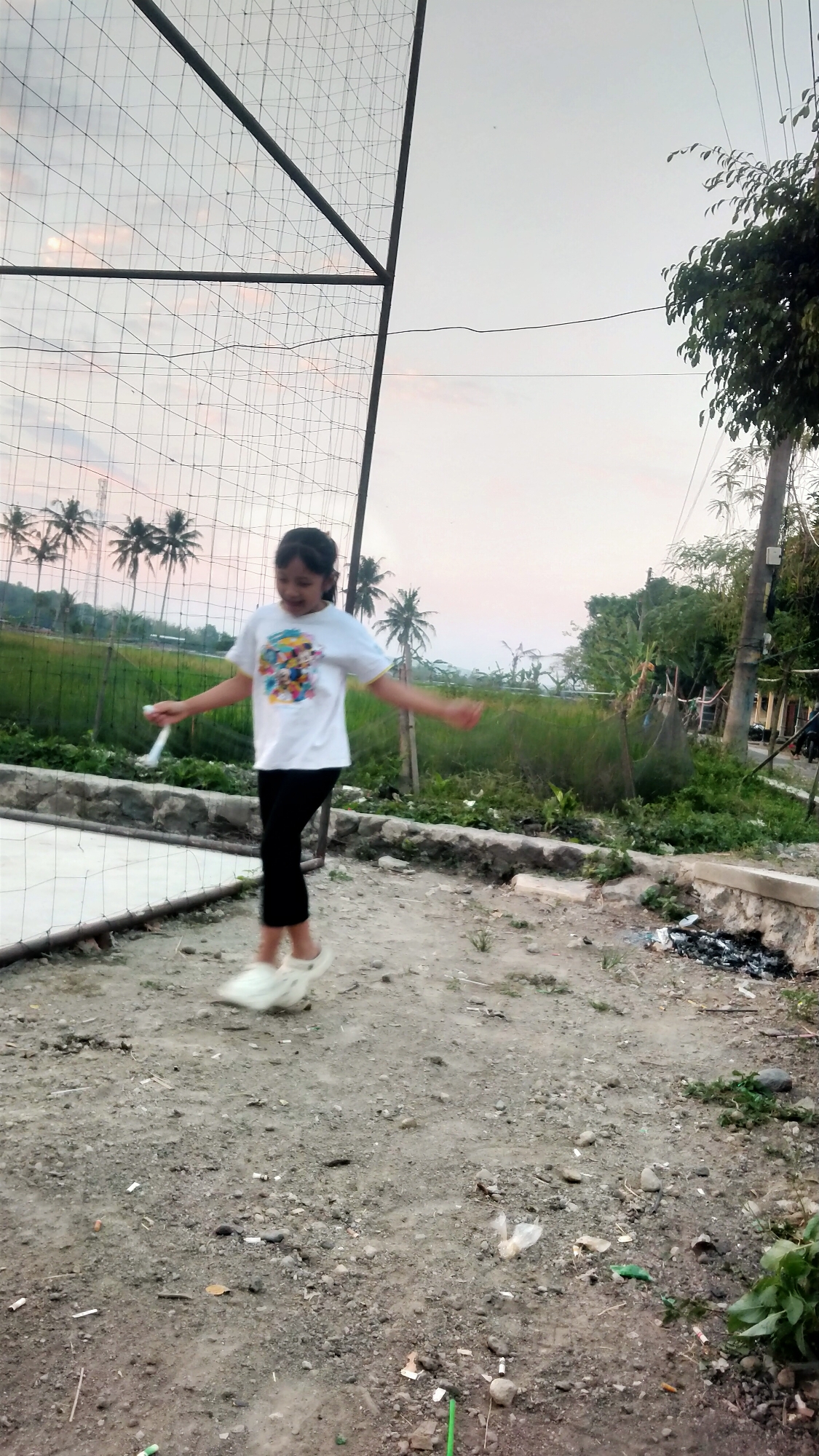An Indonesian girl in a white t-shirt and black pants is shown mid-jump rope activity in an outdoor, unpaved area. The background features a field with palm trees, suggesting a tropical or subtropical climate. A large, dark metal structure with netting is visible on the left side of the frame, possibly part of a sports court. The ground is covered with dirt and small stones, with some scattered debris including cigarette butts and plastic. A damaged concrete wall runs horizontally across the middle ground, separating the unpaved area from the grassy field. The sky is overcast with a soft, diffused light, indicative of either dawn or dusk. The scene appears to be located in a rural or semi-rural part of Ketoyan, Indonesia.