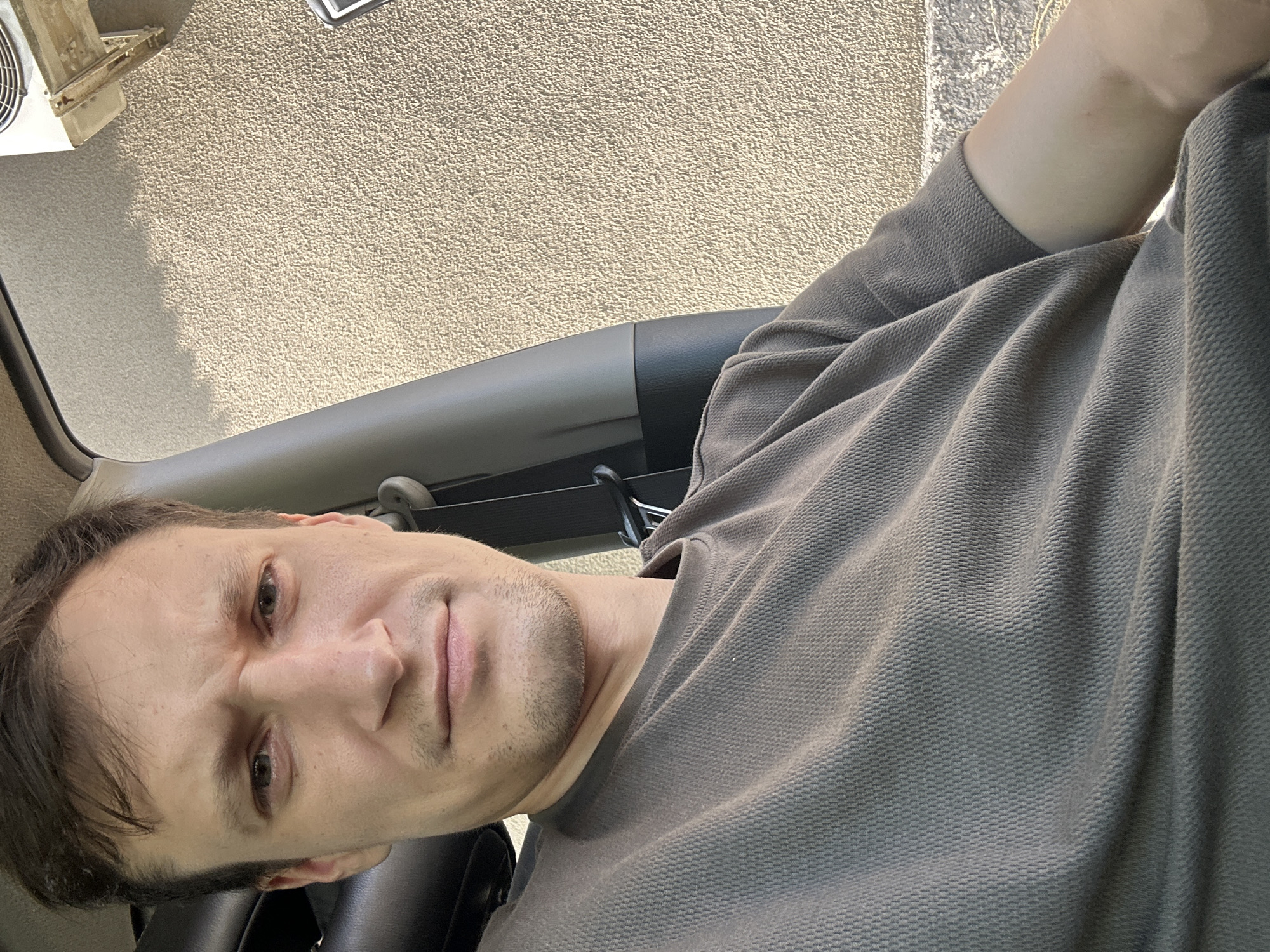 A man with short brown hair and a slight stubble is sitting in what appears to be the interior of a vehicle. He is wearing a dark olive-green t-shirt with a textured fabric. His facial expression is neutral, with furrowed brows. To his right, a textured beige wall is visible, and on the top right of the frame, part of an air conditioning unit is mounted on the wall. The background suggests an outdoor setting, possibly in the city of Shatava, Ukraine. The lighting indicates it is daytime.