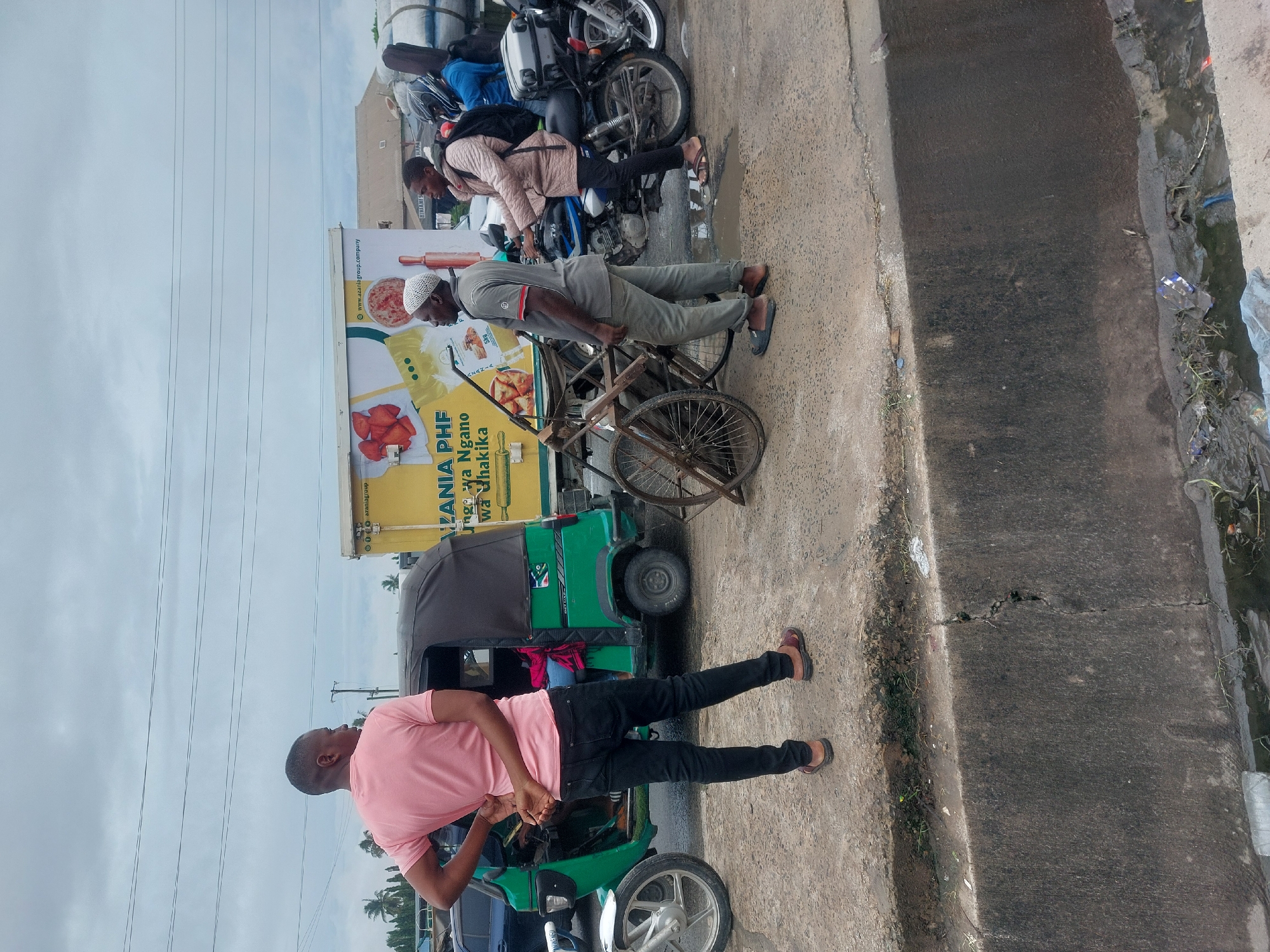 The image shows a street scene in Dar es-Salaam, Tanzania. The street is bordered by a concrete sidewalk and a drainage ditch. Several people are present. One man is standing on the sidewalk next to a green and black auto rickshaw (tuk-tuk), wearing a pink t-shirt and black jeans. Another man, wearing grey clothing and a white cap, is next to a bicycle with a rusted frame and wheels. Another person, wearing a light brown jacket and black pants, is seated on a parked motorcycle. A vehicle is parked behind the auto rickshaw. A large advertising sign with the text "TANZANIA PHF... Ngano wa uhakika" is visible on a yellow vehicle with a green canopy.
