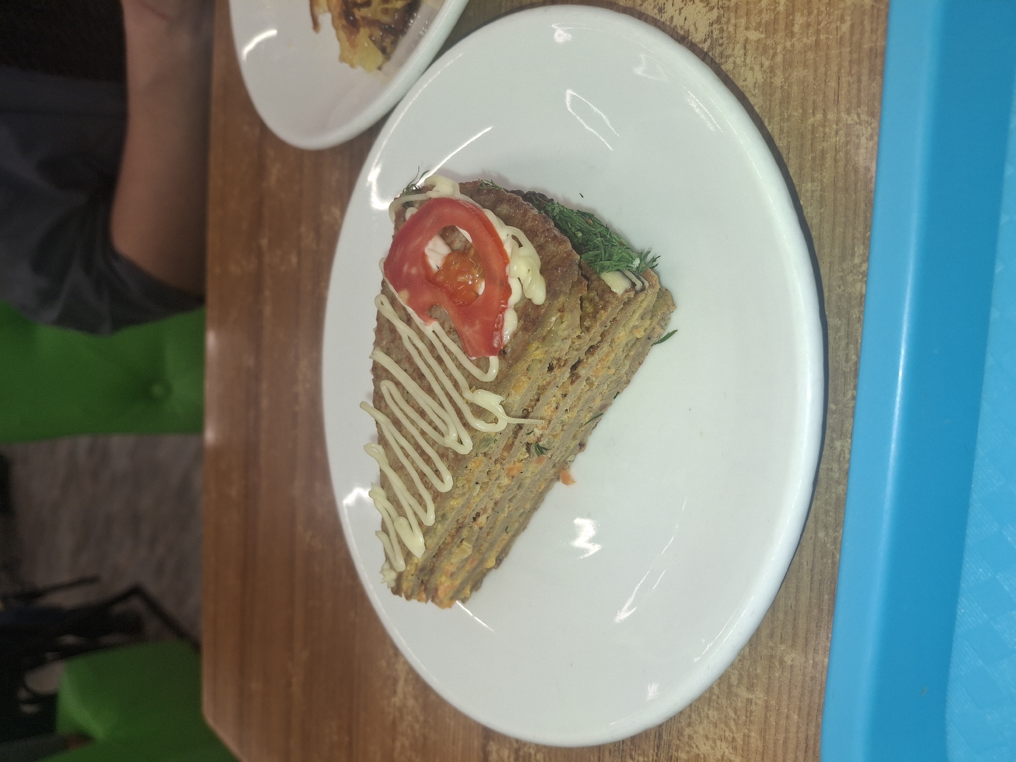Here is a forensic description of the image:
A top-down, medium shot shows a slice of layered food item, possibly a cake or savory pie, on a white plate. The slice is triangular and appears to be composed of multiple thin layers with filling in between. It is topped with a slice of tomato, a zigzag pattern of white sauce, and some green herbs, likely dill. The plate rests on a wooden table surface. To the right of the plate is a blue plastic tray. A second plate, with what looks like another piece of food, is visible in the upper left corner. A person’s arm and portion of their clothing are visible in the upper left corner, adjacent to a green chair. The scene is brightly lit, suggesting it is indoors. No specific identifying information regarding Almaty, Kazakhstan, is discernible from the image.
