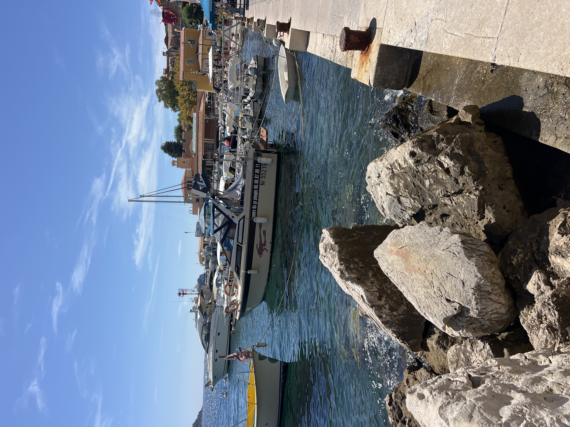 The image captures a sunny day in the port of Villefranche-sur-Mer, France. The foreground features a rocky shoreline with large, light-colored boulders. The water is a deep blue with shimmering sunlight reflecting on its surface. Several boats are moored in the harbor, including a white yacht with a striped awning, a smaller white boat with a dark canopy, and a yellow vessel. In the background, buildings with terracotta roofs and ochre-colored facades line the waterfront, suggesting a Mediterranean architectural style. The sky is bright blue with scattered white clouds. The scene is tranquil and picturesque, typical of a French Riviera harbor.