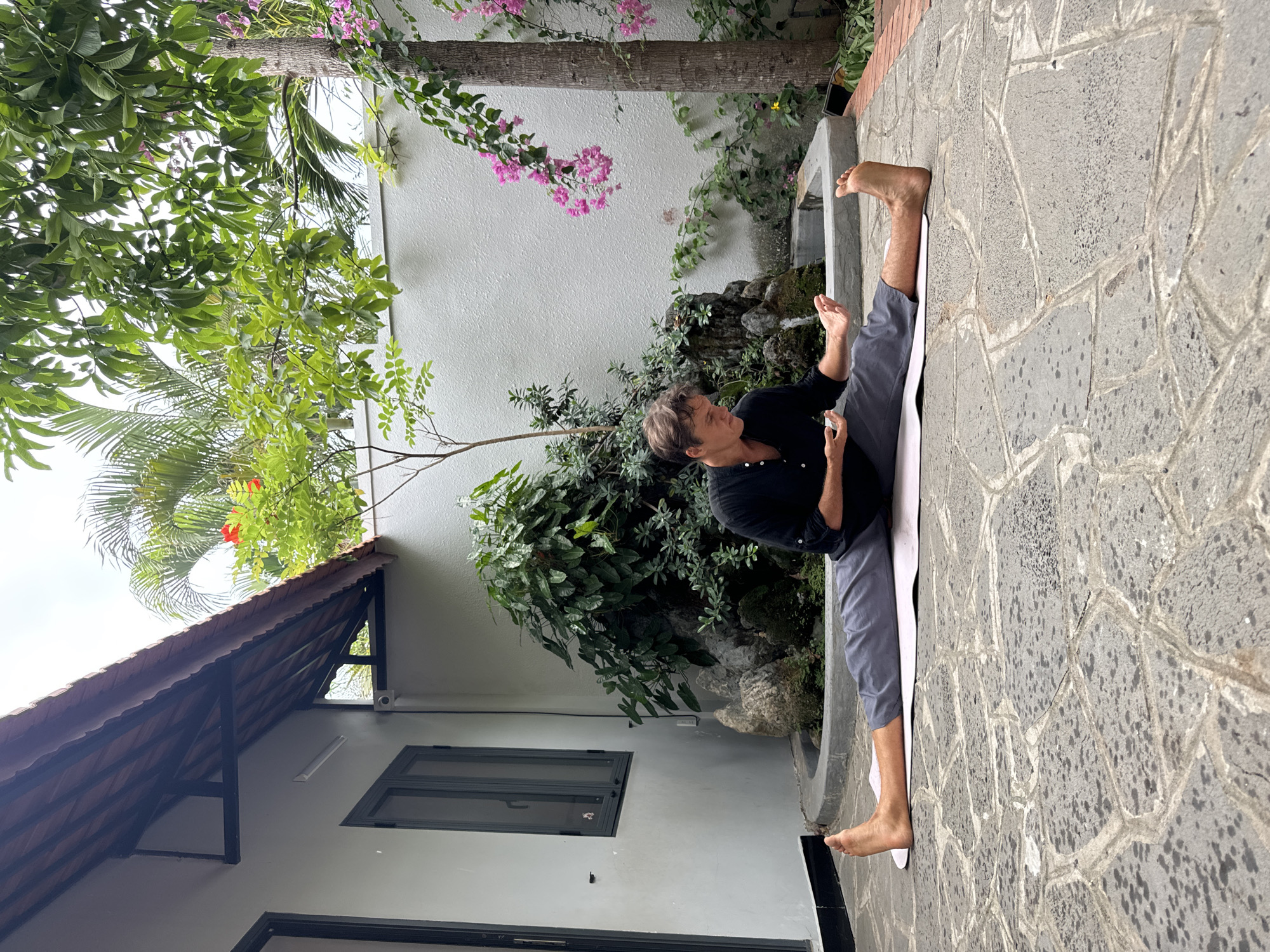 An image shows a Caucasian man seated in a wide straddle pose (likely doing yoga) on a white mat on a gray stone terrace. He is wearing a dark long-sleeved shirt and gray pants. He faces forward, gesturing with his hands. Behind him is a white wall with green foliage, a small fountain feature, and pink flowers growing on a tree branch above. To the left is a building with a dark roof overhang and windows. The scene appears to be in an outdoor courtyard or garden setting, likely in Mũi Né, Vietnam.