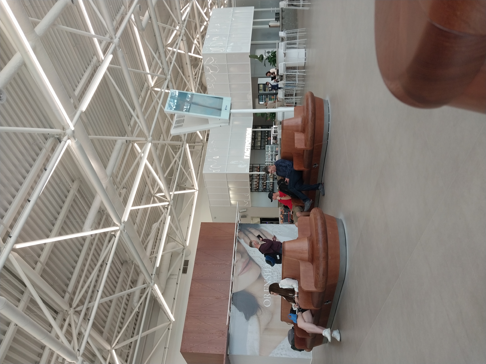 The image depicts the interior of an airport or a similar public transportation hub, likely located in Orenburg, Russia, as indicated by a large advertisement featuring the text "ORENSHA" which is a prominent local brand. The scene shows a waiting area with modern, curved, brown leather seating arrangements. Several individuals are seated or standing, engaged in various activities such as looking at their phones, reading, or conversing.
In the foreground, a woman in a white shirt and black shorts is seated on the left side of a curved bench, with her legs crossed. To her right, another woman in a white shirt is seated, her backpack on the floor beside her. A man wearing a burgundy shirt and jeans stands near the center of the frame, holding a phone and a book.
In the middle ground, a similar curved bench accommodates two men. One man in a red shirt and dark pants is seated, while another man in a dark blue shirt and jeans is seated and looking at his phone. Further back, a modern cafe or restaurant area is visible with white counters, high stools, and bar seating. People are seen interacting in this area, including a server behind the counter. Above the cafe, a large digital display screen is mounted on a white stand.
The environment is characterized by a high, industrial-style ceiling with exposed white beams and fluorescent lighting. The floor is a light-colored tiled surface. The overall aesthetic is contemporary and functional, with elements of wood and leather contributing to a sense of comfort in the waiting area. A large advertisement featuring a woman's face and the text "ORENSHA E.1939" is prominently displayed on a wall in the background, further suggesting a connection to Orenburg.