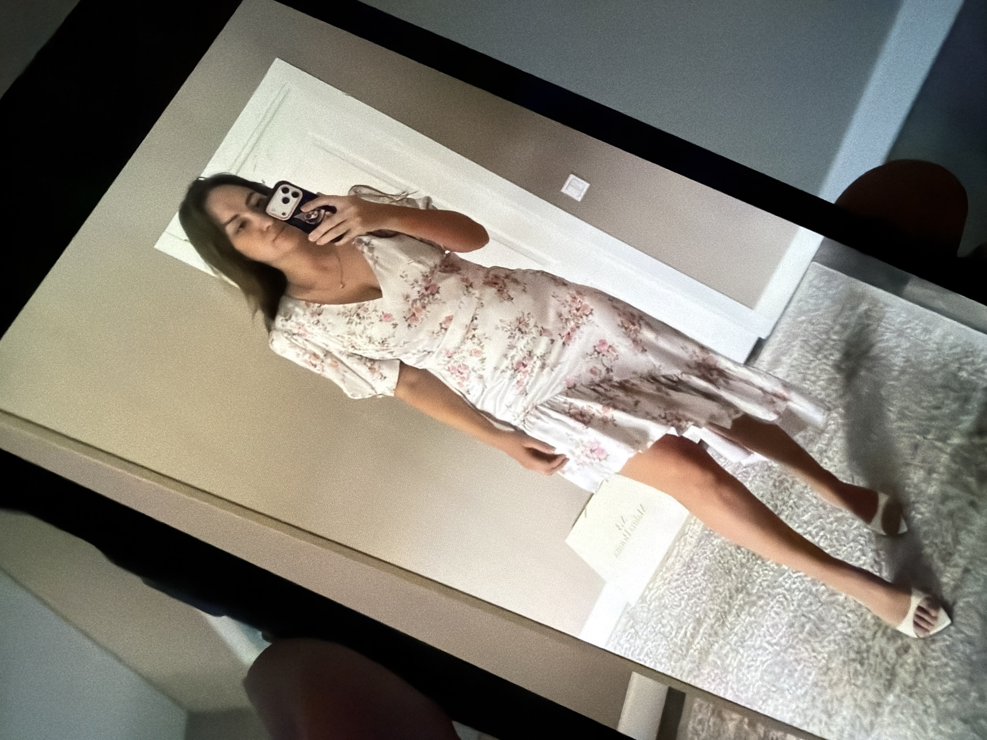 The image shows a person taking a mirror selfie with a phone. The person is wearing a floral print dress and white pointed-toe heels. They are standing on a textured rug in a room. There is a door in the background. The image is taken from an angled perspective, showing the phone screen reflecting the scene. There is no identifiable information in the image to suggest a location in Malinovka, Russia.