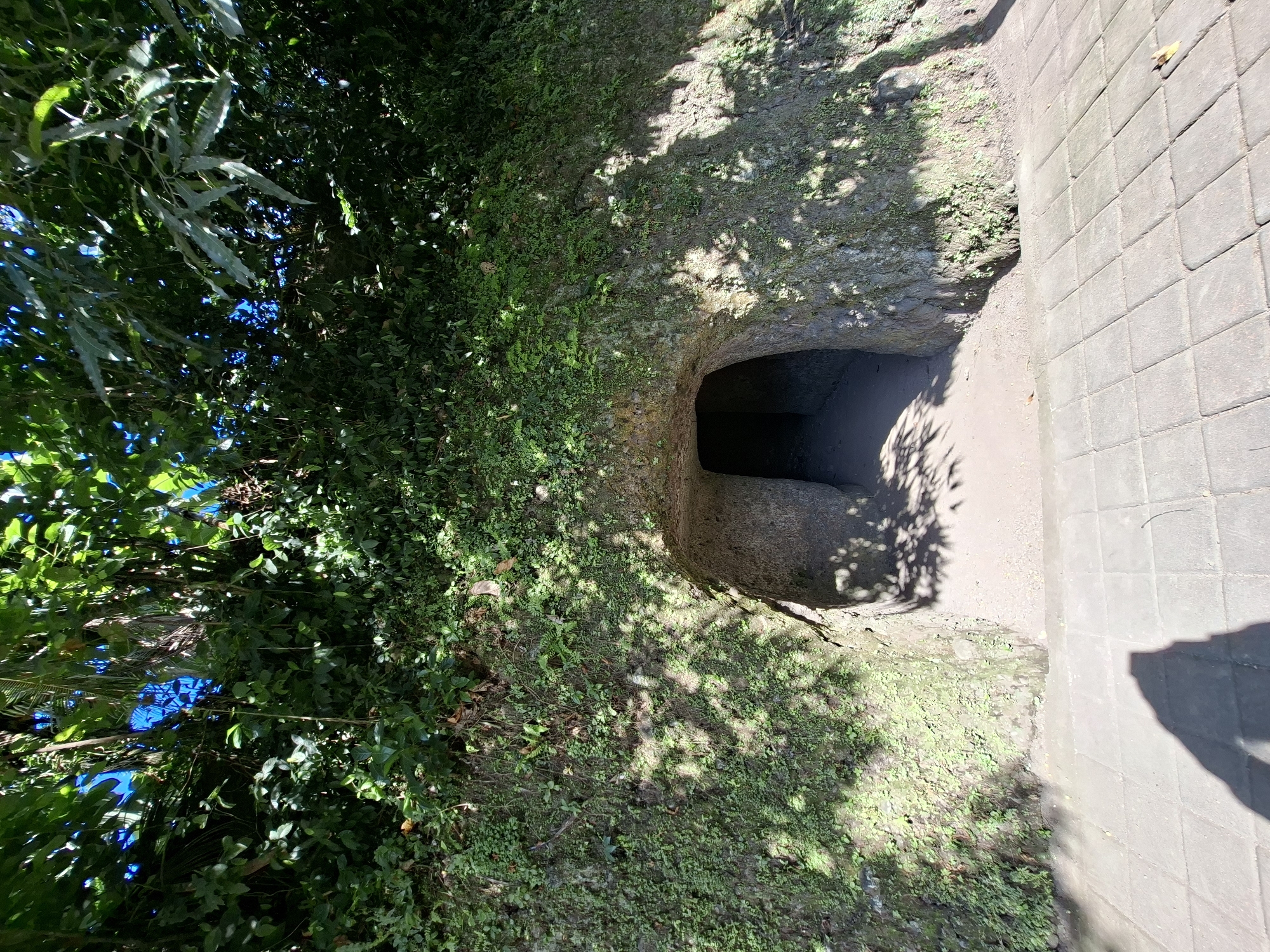 The image shows the entrance to a tunnel carved into a moss-covered hillside. The tunnel entrance is roughly arched and appears to be made of rough-hewn stone. The hillside is covered in lush green vegetation, with sunlight filtering through the leaves of trees above, casting dappled shadows on the ground and the tunnel. The ground in front of the tunnel is paved with interlocking gray stones, and a long shadow of a person is visible on the pavement in the foreground. The tunnel entrance is dark, suggesting depth within. The overall scene is one of a natural, perhaps ancient, structure integrated into a verdant landscape. This entrance is located at Goa Gajah, a significant archaeological site in the region of Banjarangkan, Indonesia.