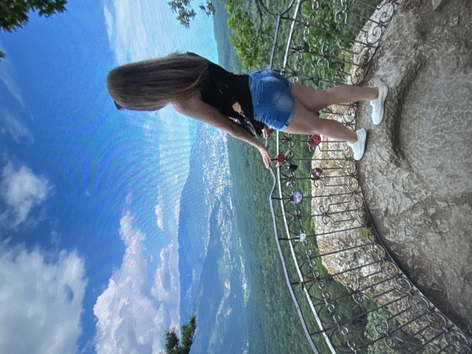 A person stands on a stone platform behind a decorative metal railing, looking out at a vast landscape. The person is wearing a black tank top and denim shorts, with white sneakers on their feet. Their hair is long and blonde, falling down their back. The railing is adorned with several heart-shaped locks, likely a symbolic gesture of love.
The platform overlooks a valley filled with lush green trees, which recede into rolling hills and mountains in the distance. A city or town is visible nestled within the valley, with buildings scattered amongst the greenery. The sky above is a bright blue, dotted with fluffy white clouds. The overall scene suggests a scenic viewpoint, possibly a tourist attraction or a natural landmark. The quality of the image is moderate, with a slightly grainy texture and visible pixelation, which may indicate it was taken with a lower-resolution camera or has been digitally altered.