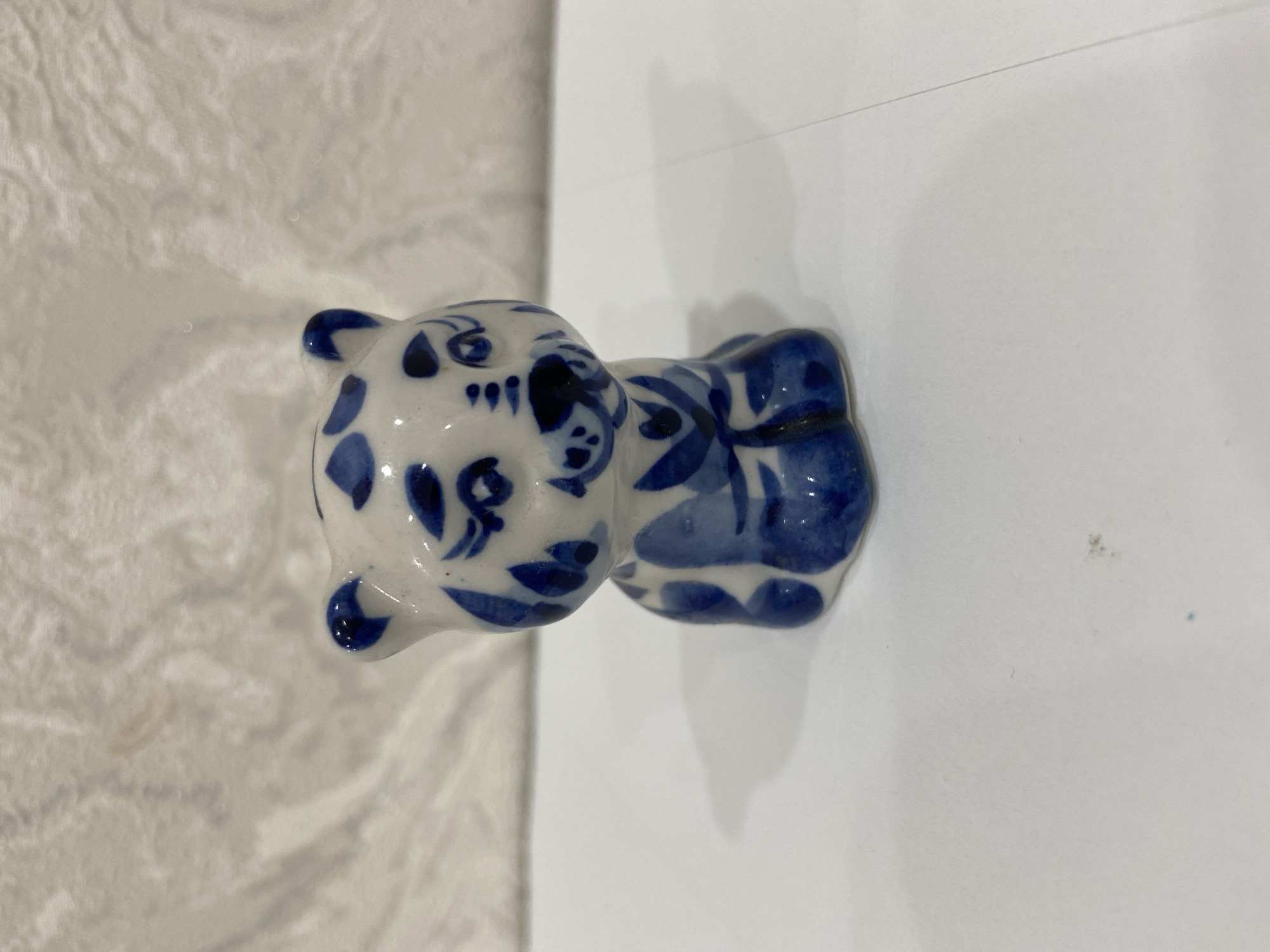 The image shows a small, ceramic figurine of a tiger, painted with blue and white patterns. The figurine is sitting on a white surface, which appears to be a table or shelf. Behind the figurine, there is a textured white background. The lighting is soft and diffused, casting a gentle shadow to the left of the figurine. The figurine is centrally located in the frame. There is no information in the image to suggest its location in Balashikha, Russia.