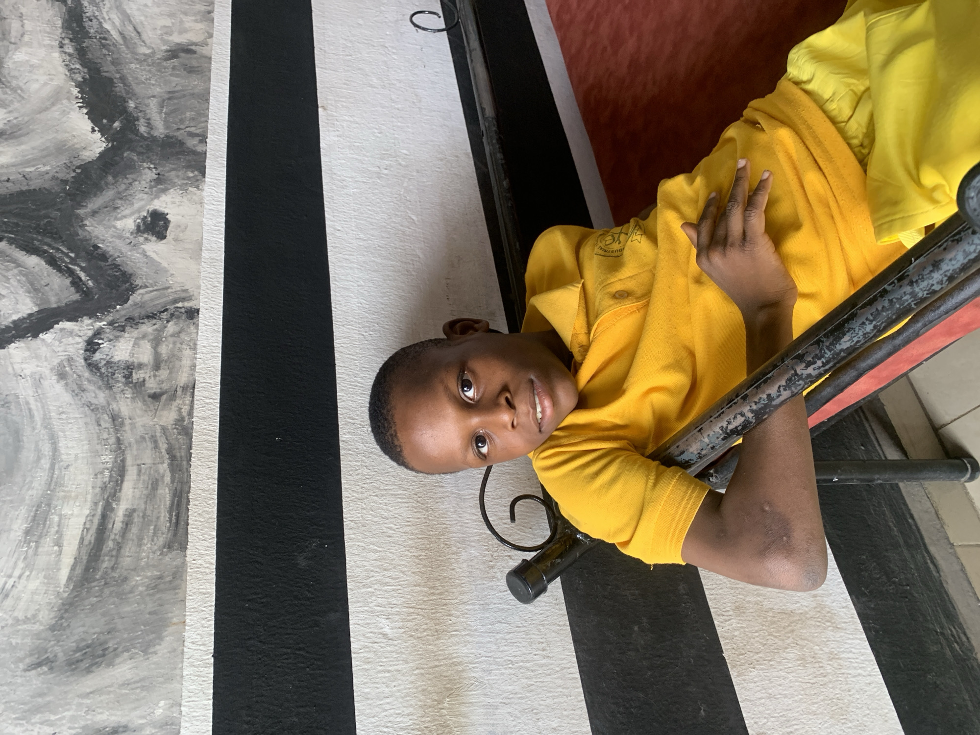 A child wearing a bright yellow shirt is lying on a metal-framed surface with a red cushion, indoors. The background consists of black and white stripes and a gray, textured surface resembling a painted wall. The location is presumed to be Jos, Nigeria.