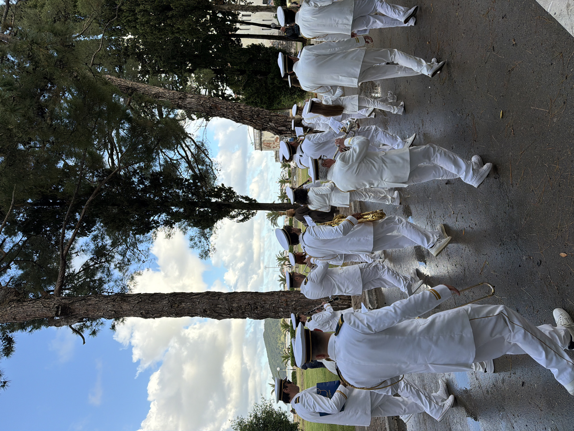 The image shows a procession of people dressed in white uniforms and white hats. They appear to be part of a marching band, as some are holding and playing musical instruments like trombones, saxophones, and clarinets. The procession is taking place on a paved surface, likely a street or walkway. There are trees to the left of the group. In the background, there is a view of a partly cloudy sky, mountains, and palm trees. Based on the foliage and architecture visible in the background, the location is likely in Budva, Montenegro.