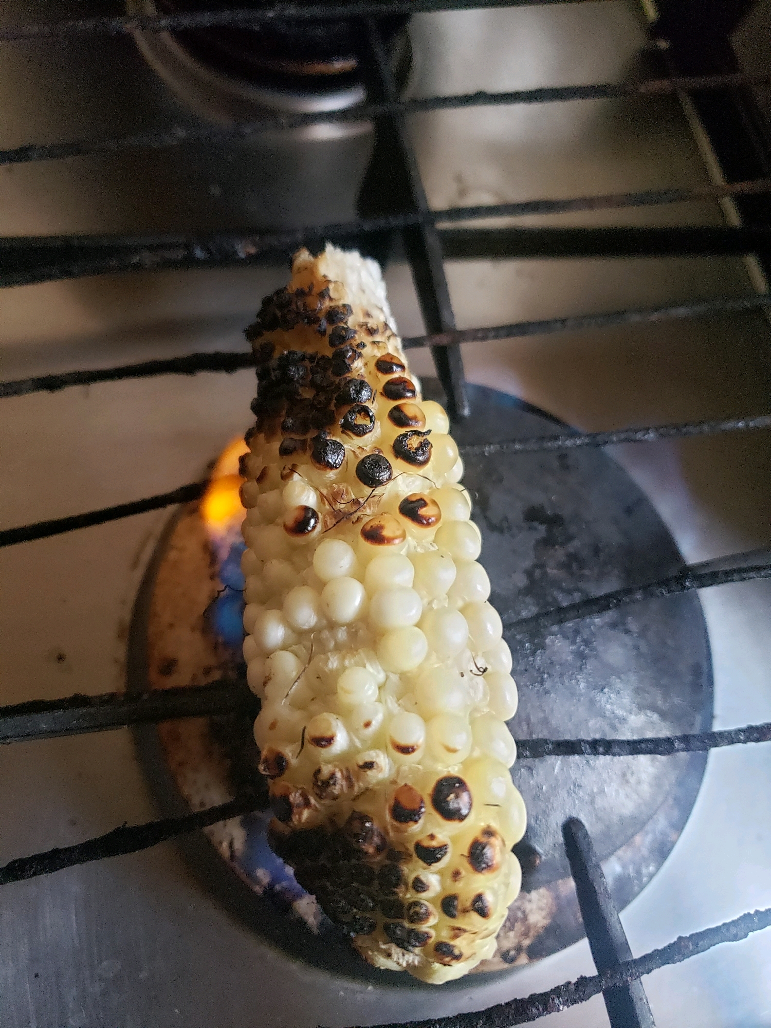 A close-up, overhead shot shows a partially shucked ear of corn being roasted directly over a gas burner. The corn, with its kernels exposed, has begun to char in places, with some kernels showing dark brown or black spots. The flame from the burner is visible, licking the underside of the corn. The corn rests on a metal grate, which is part of a gas stove. The environment appears to be a kitchen.