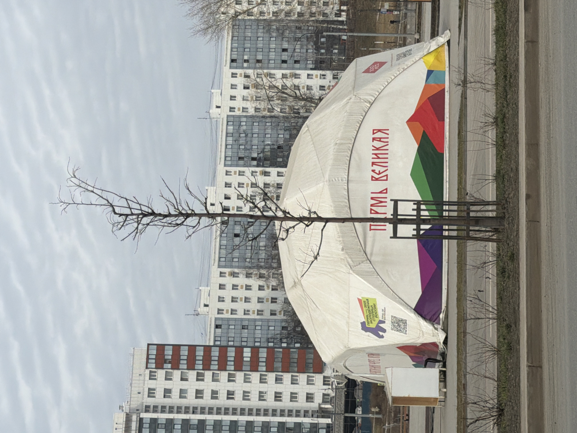 A slightly high-angle, outdoor shot captures a city street in Perm, Russia. A barren tree stands prominently in the foreground, its leafless branches reaching across the scene. Behind the tree is a large white tent with colorful graphics and Cyrillic text reading "ПЕРМЬ ВЕЛИКАЯ". The tent sits on the edge of a road and extends back toward a backdrop of high-rise buildings with multiple windows. The sky is overcast, contributing to the muted color palette. A concrete barrier separates the tent from the street. The light is diffused, creating soft shadows.