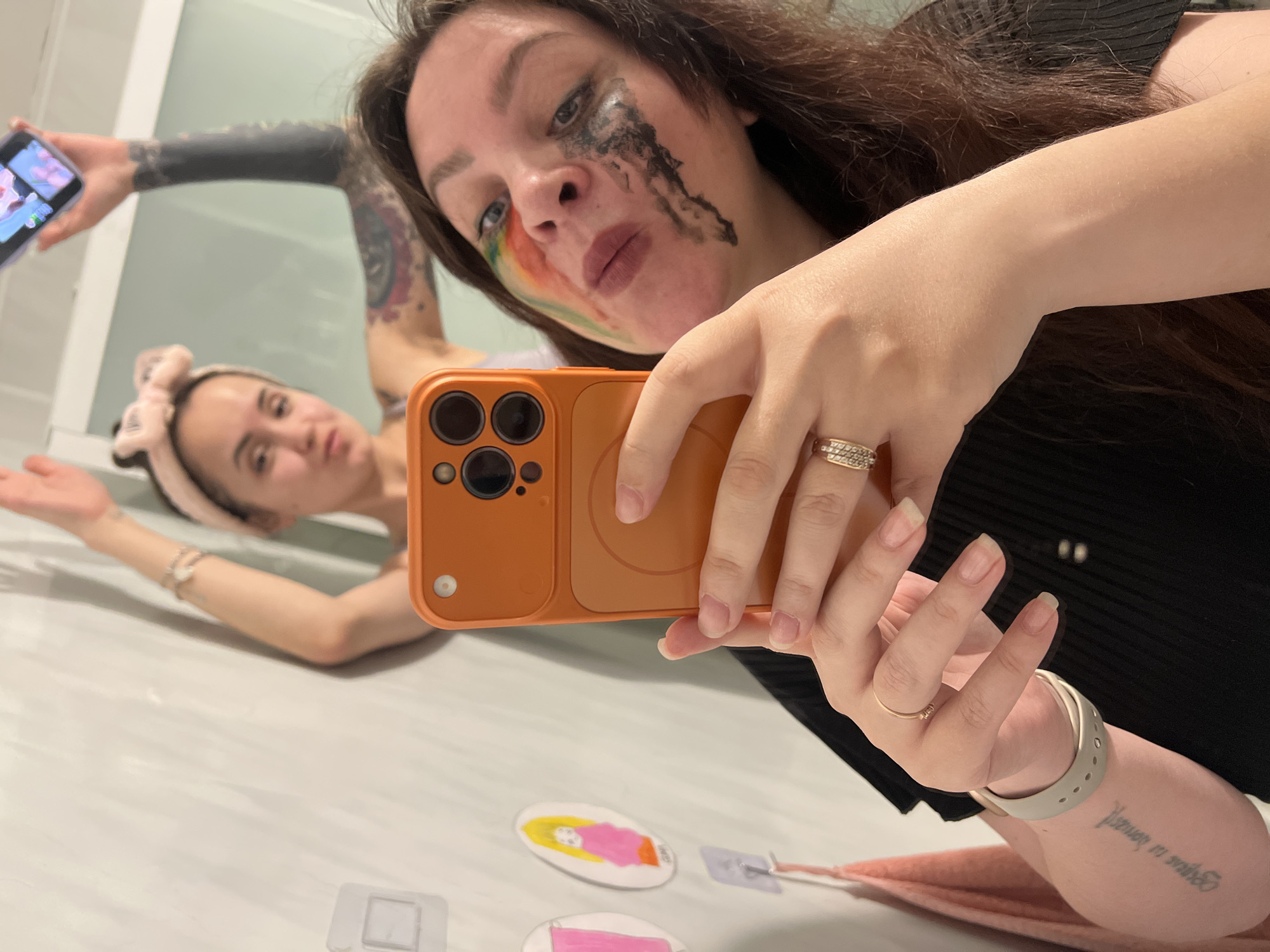 A selfie taken in a bathroom or similar setting. A woman is holding a phone with an orange case, taking the picture. She has makeup smeared on her face, including a rainbow pattern on her cheek and black streaks under her eye. Another woman is visible in the background, lying down and looking at the camera. The background appears to be a white or light-colored surface. There are small drawn portraits on the surface. One of the woman's arms has a tattoo on it. The location in the city of Noviy, Russia, is unknown.