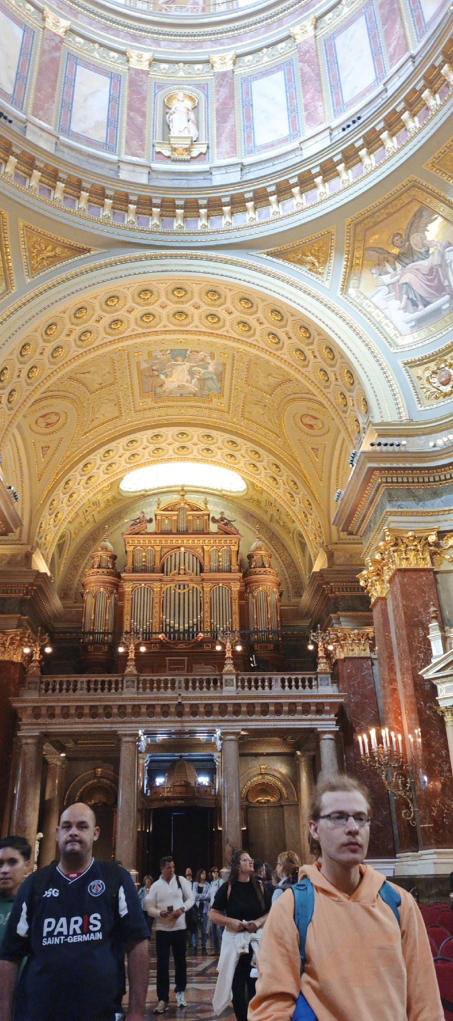 This photograph, taken indoors, appears to be in St. Stephen's Basilica in Budapest, Hungary. The image focuses on the basilica's ornate, high-ceilinged interior, featuring a large pipe organ positioned high above, and a complex architectural design embellished with gold accents, paintings, and statuary.
In the foreground, several individuals are seen walking and standing. The lighting is predominantly natural, filtering through the high windows and contributing to the warm tones of the basilica's interior. The scene captures a blend of architectural grandeur and human activity.