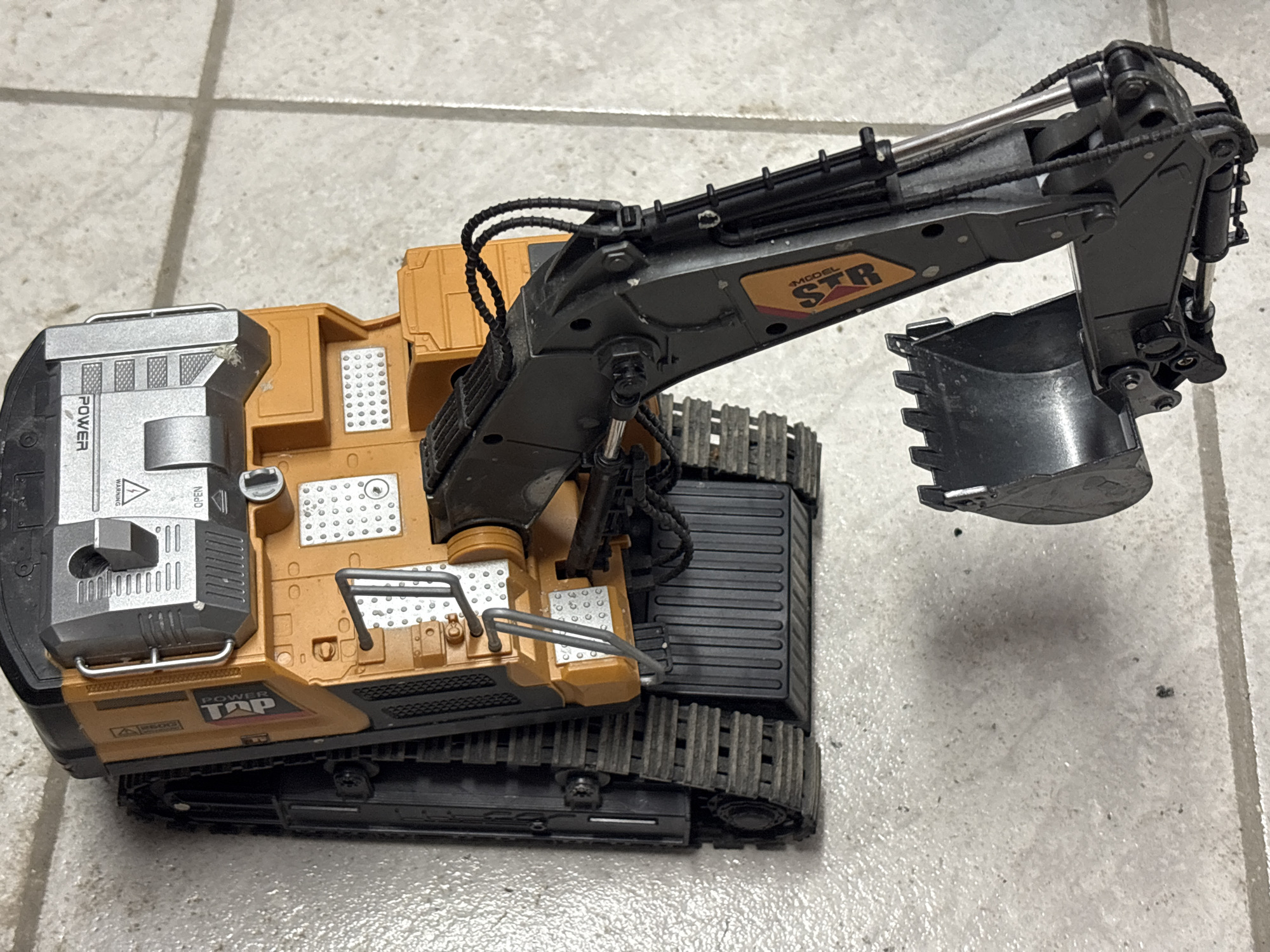 The image depicts a toy excavator resting on a tiled floor. The excavator is primarily black and orange, with a silver panel on the left side. The boom and bucket are extended upwards. The tiles appear to be light gray or white with darker grout lines. There is no indication of the location being Plovdiv, Bulgaria, or any specific city, nor any indication of people or actions beyond the presence of the toy.