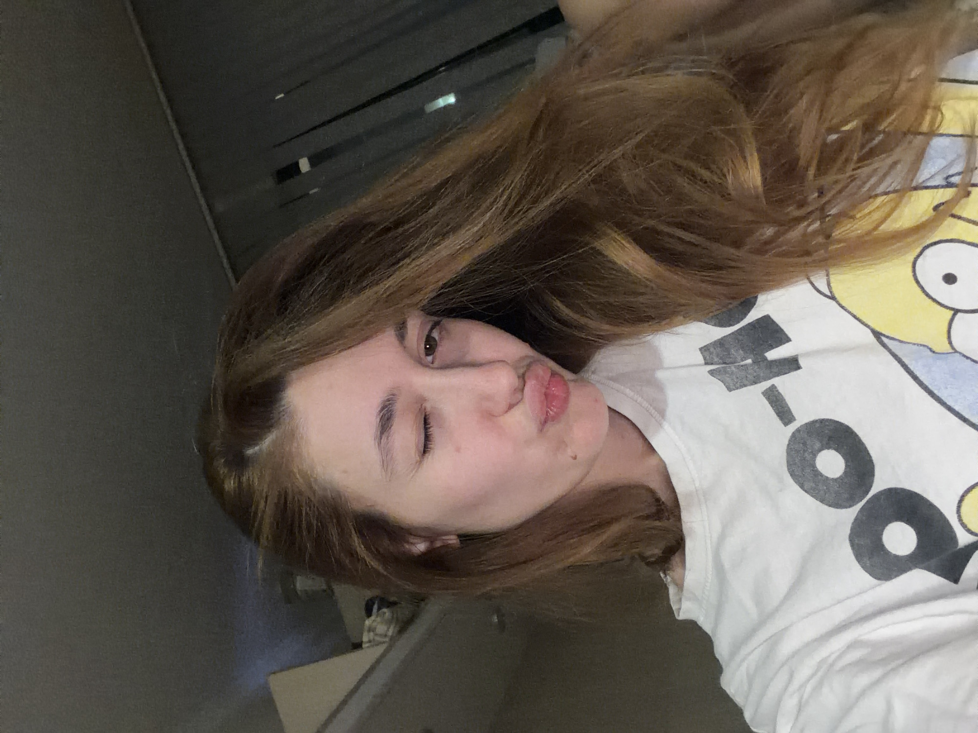 A young woman with brown hair is pictured indoors. She is wearing a white t-shirt featuring a cartoon character and making a kissing face with one eye closed. The background includes a dark wall or surface. There is insufficient information to determine a specific location in Sofia, Bulgaria.