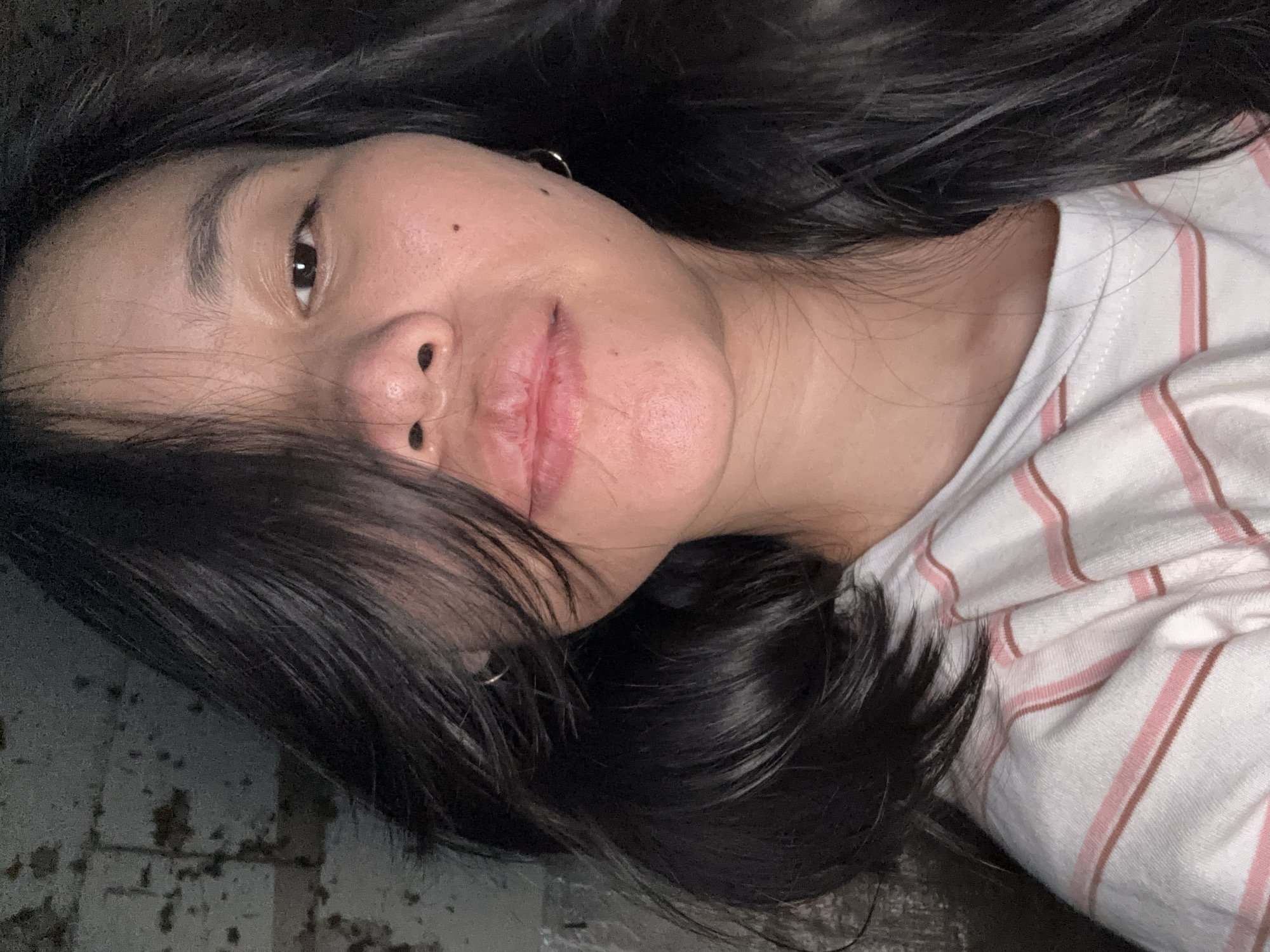 The image shows a close-up, selfie-style portrait of a young East Asian woman. She has dark, wavy hair that partially obscures her face, with strands falling across her forehead and cheek. Her skin is fair with some visible texture and a few small moles. Her right eye is visible, and she has a subtle expression. Her lips are slightly parted. She is wearing a white t-shirt with thin pink and brown stripes. The background appears to be a distressed, textured wall with a gritty, industrial feel. The lighting is dim and somewhat harsh, creating shadows on her face and the background. The overall scene suggests an indoor setting, possibly an urban environment given the background, but no specific location in East Jakarta, Indonesia can be determined from the visual information provided.