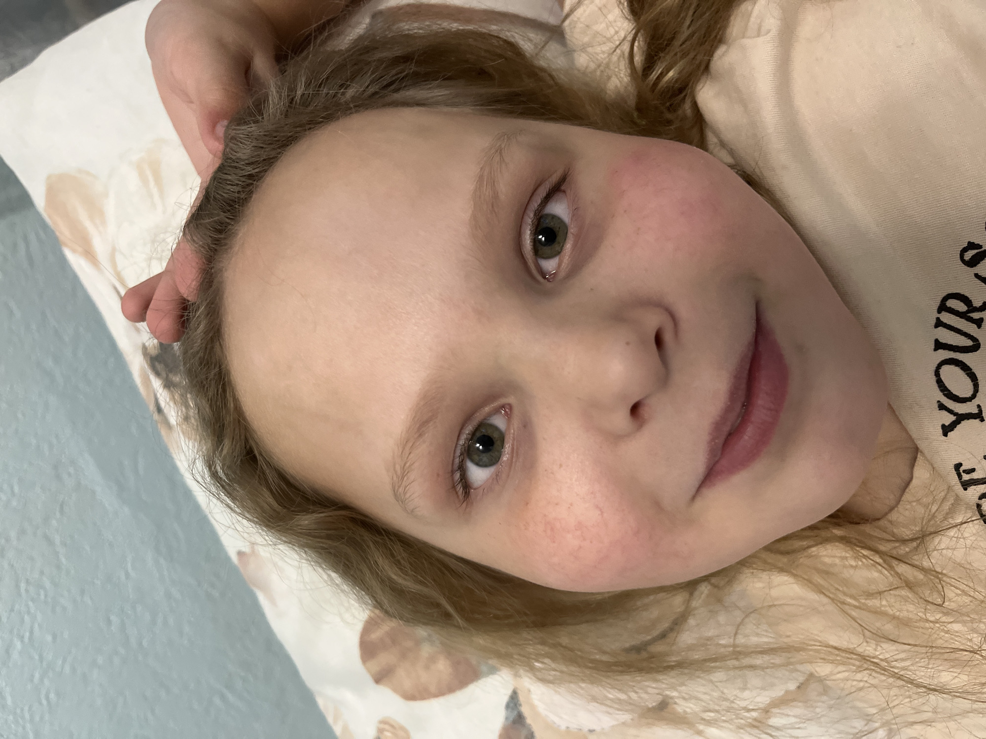 A young girl with blond hair and green eyes is lying down, looking up at the camera. She has a slight smile. Her skin is pale with reddish cheeks. Her right hand is positioned near her head on a pillow. Part of a beige t-shirt with partial lettering is visible near her neck. The pillowcase beneath her has a beige and white pattern. The background appears to be a light blue wall or surface. The location in the city of Izhevsk, Russia, cannot be determined from the image.