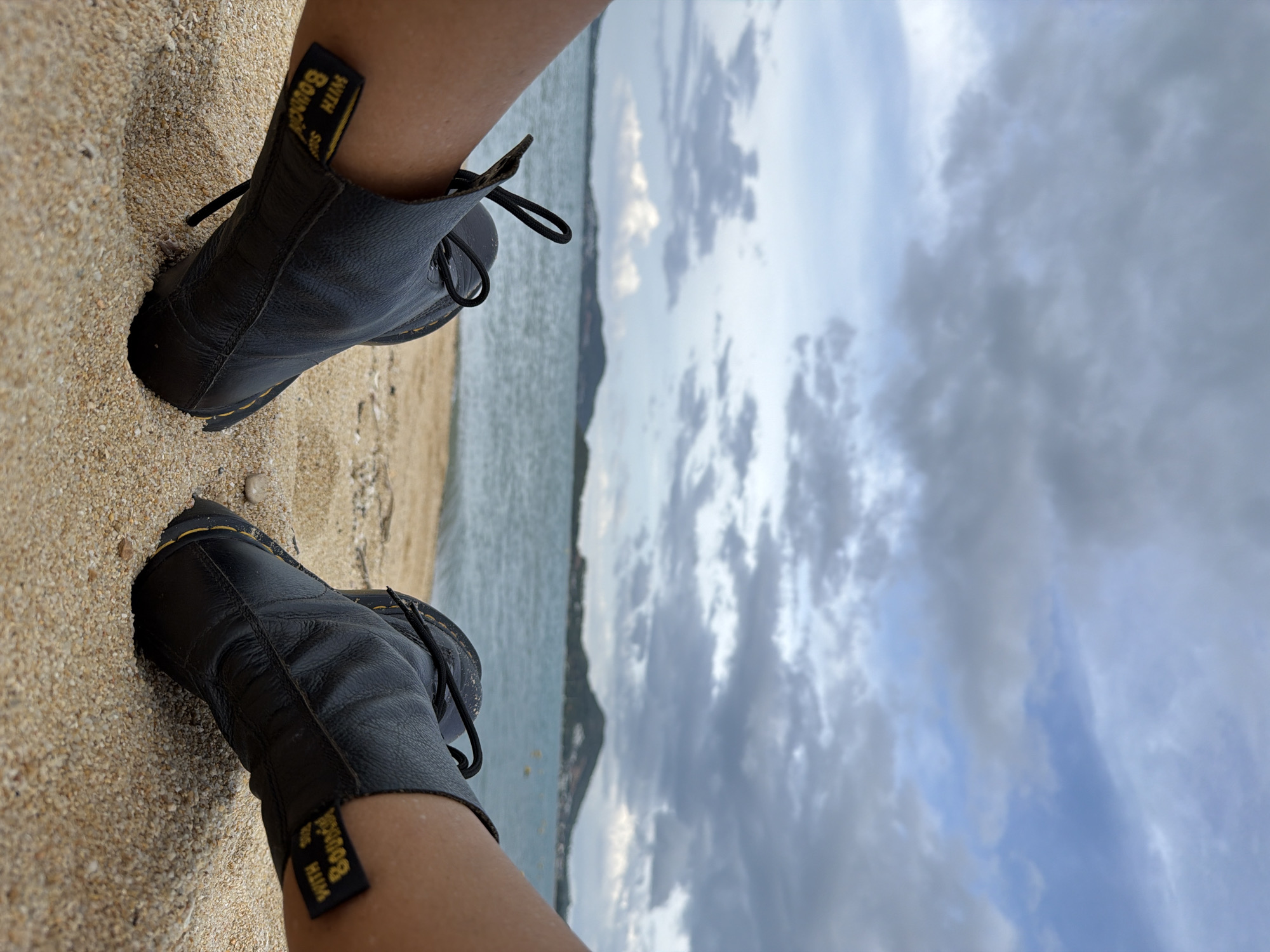The image shows a first-person perspective of someone wearing black leather boots with yellow stitching, with their feet partially submerged in sand on a beach. In the background, there is a body of water, likely the sea, with a distant shoreline featuring hills or mountains. The sky is cloudy, with a mix of gray and blue tones. The specific location is the beach in Ko Samui District, Thailand. The boots appear to be Dr. Martens, indicated by the yellow pull tabs with black lettering that read "WITH SOLES". The sand is light brown and appears wet in some areas closer to the water. The overall scene suggests a relaxed or contemplative moment at the seaside.