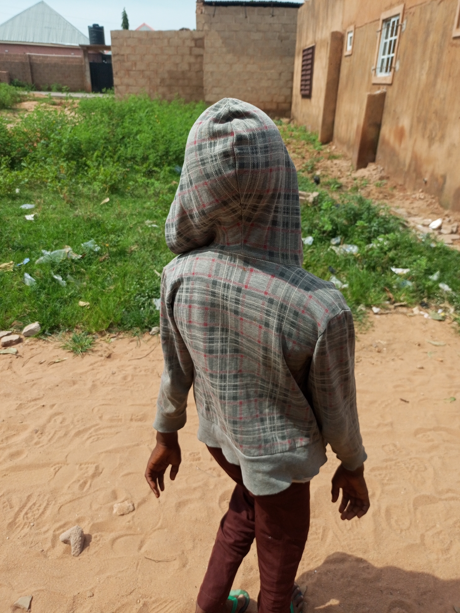 A person wearing a gray and red plaid hooded jacket and dark red pants walks away from the camera on a sandy path. The person's left hand is extended, with the palm facing down. In the background, there is a building with an orange facade and windows, a brick wall, and green vegetation littered with plastic waste. A metal roof structure is visible on the left. The sandy ground shows footprints and scattered stones. The scene is outdoors in what appears to be a developing area.