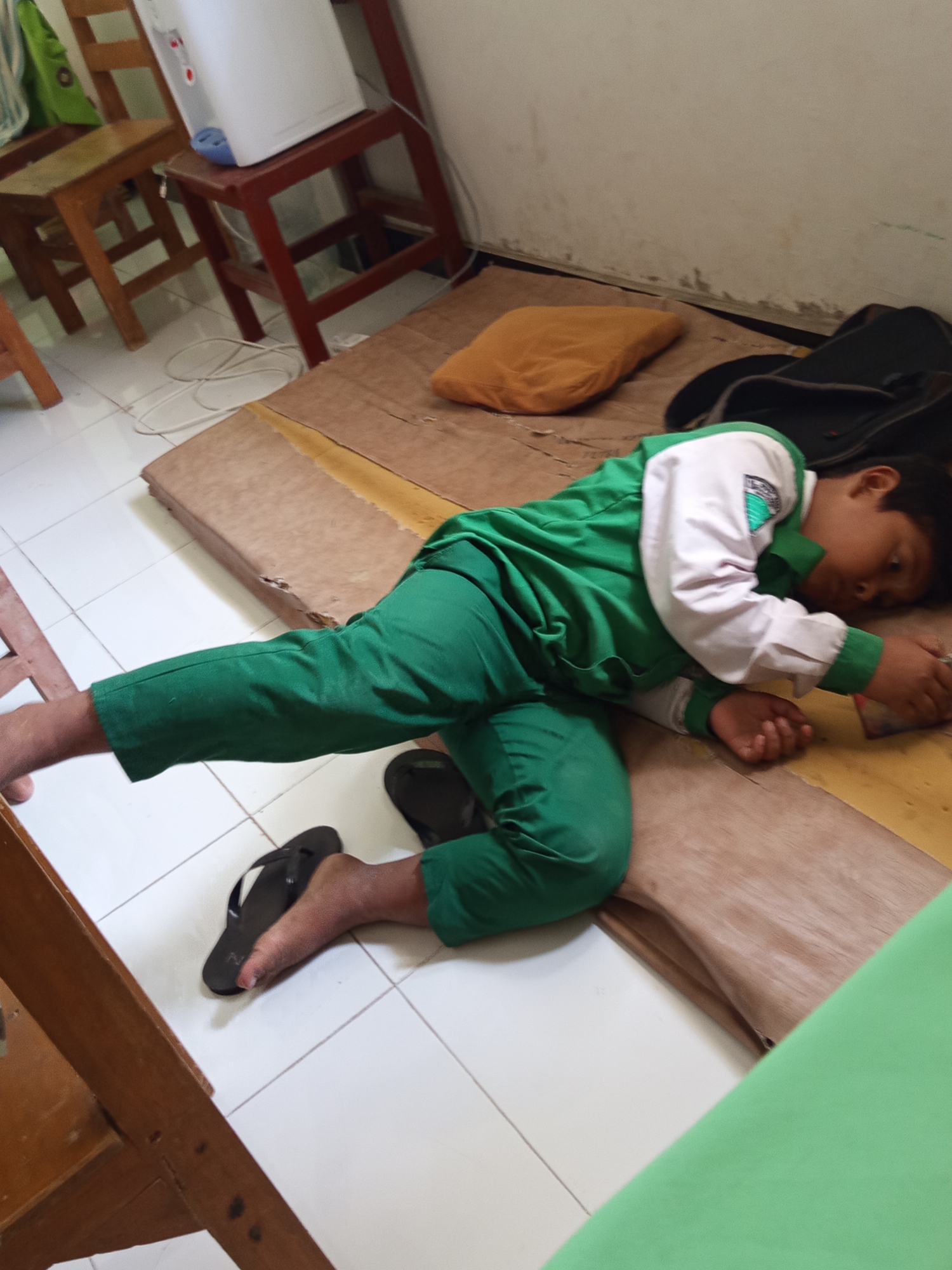 A young boy wearing a green and white school uniform and black flip-flops is lying on his side on a wooden mat on the floor in a room that appears to be a classroom or a similar educational setting. The room has white tiled flooring and a plain wall. In the background, there are wooden chairs and what looks like a white water dispenser on a wooden stool. A yellow cushion is placed near the boy. The boy is on his right side, with his head resting on his right arm, and he is looking downwards. His left leg is bent, and his right leg is extended.