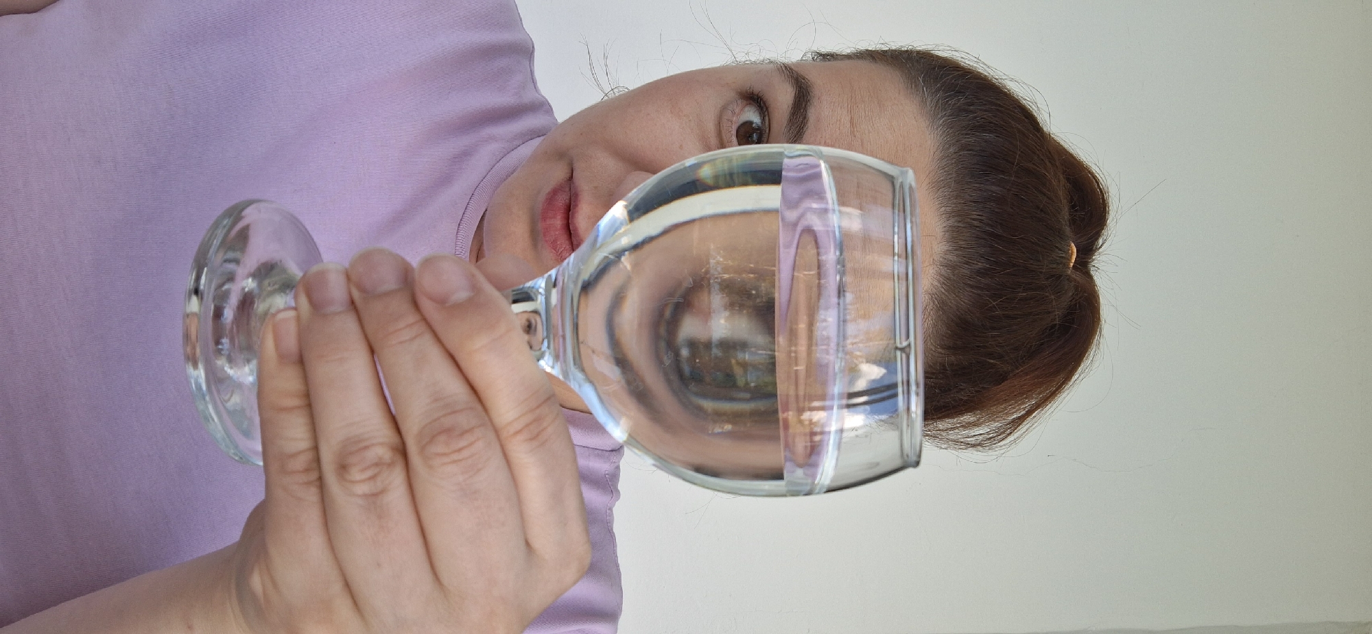 A woman with brown hair pulled back in a bun is holding a clear wine glass filled with water in front of her face. She is wearing a light purple shirt. The background is a plain white wall. The image appears to be taken indoors. No information can be gathered about Chaikovsky, Russia from the image.