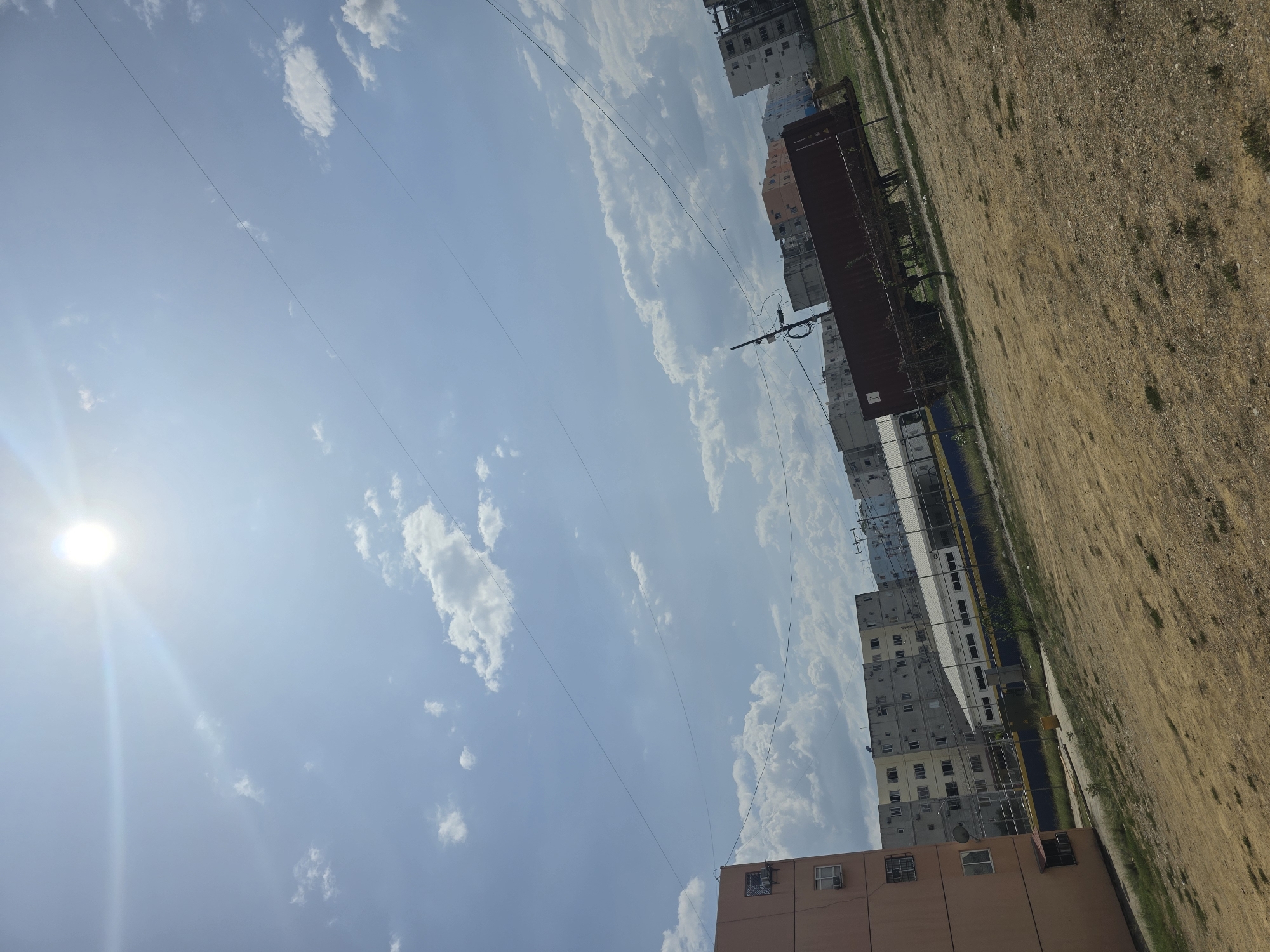 The image captures a daytime scene in Parroquia Ocumare, Venezuela. The sky is blue with scattered clouds. Power lines stretch across the sky. The foreground shows buildings, and next to the buildings is an area of dry, bare ground. In between the bare ground and the buildings appears to be a train. The sun is visible in the upper left corner, casting a bright glare.