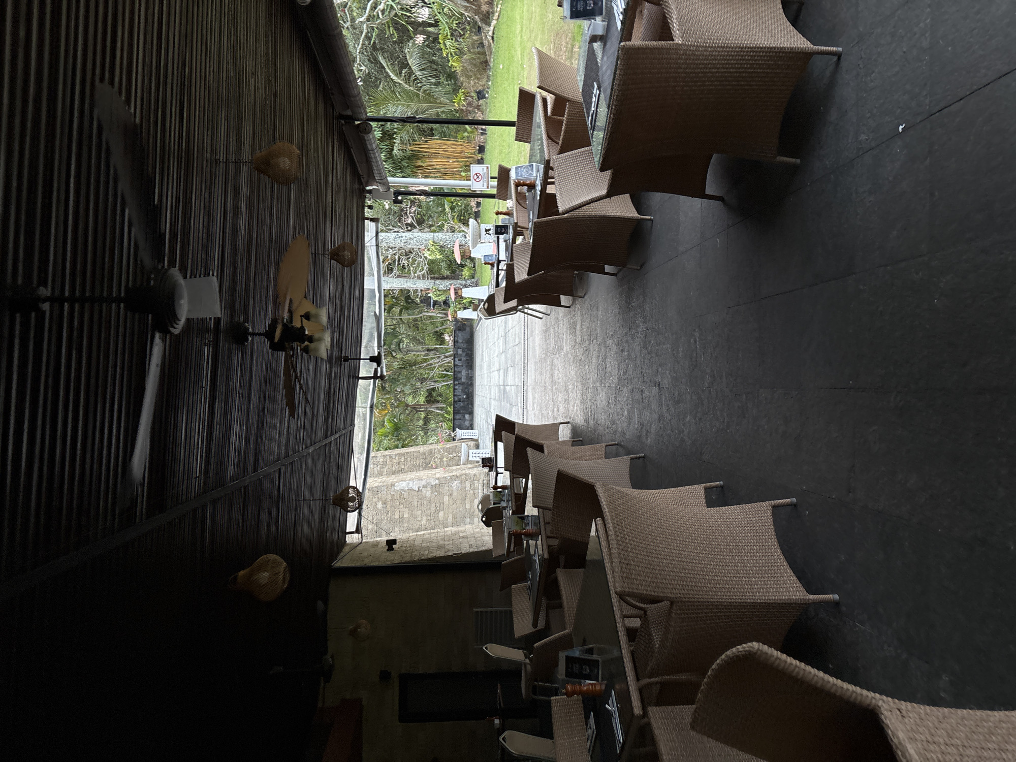 The image depicts an outdoor seating area of a restaurant or cafe, likely in Kuta Selatan, Indonesia, given the tropical foliage visible in the background and the architectural style. The scene is covered by a dark, corrugated roof supported by a dark metal frame, with several ceiling fans suspended overhead. Woven rattan chairs surround dark-topped tables, many of which are set with salt and pepper shakers and possibly menus or table tents. The floor is paved with dark, large tiles. In the background, a lush green lawn is visible, dotted with palm trees and other tropical vegetation, suggesting a warm climate. A partially visible stone wall structure stands to the left, and a doorway is also present. The lighting is dim, indicating it might be early morning, late afternoon, or an overcast day. There are no people visible in the scene.