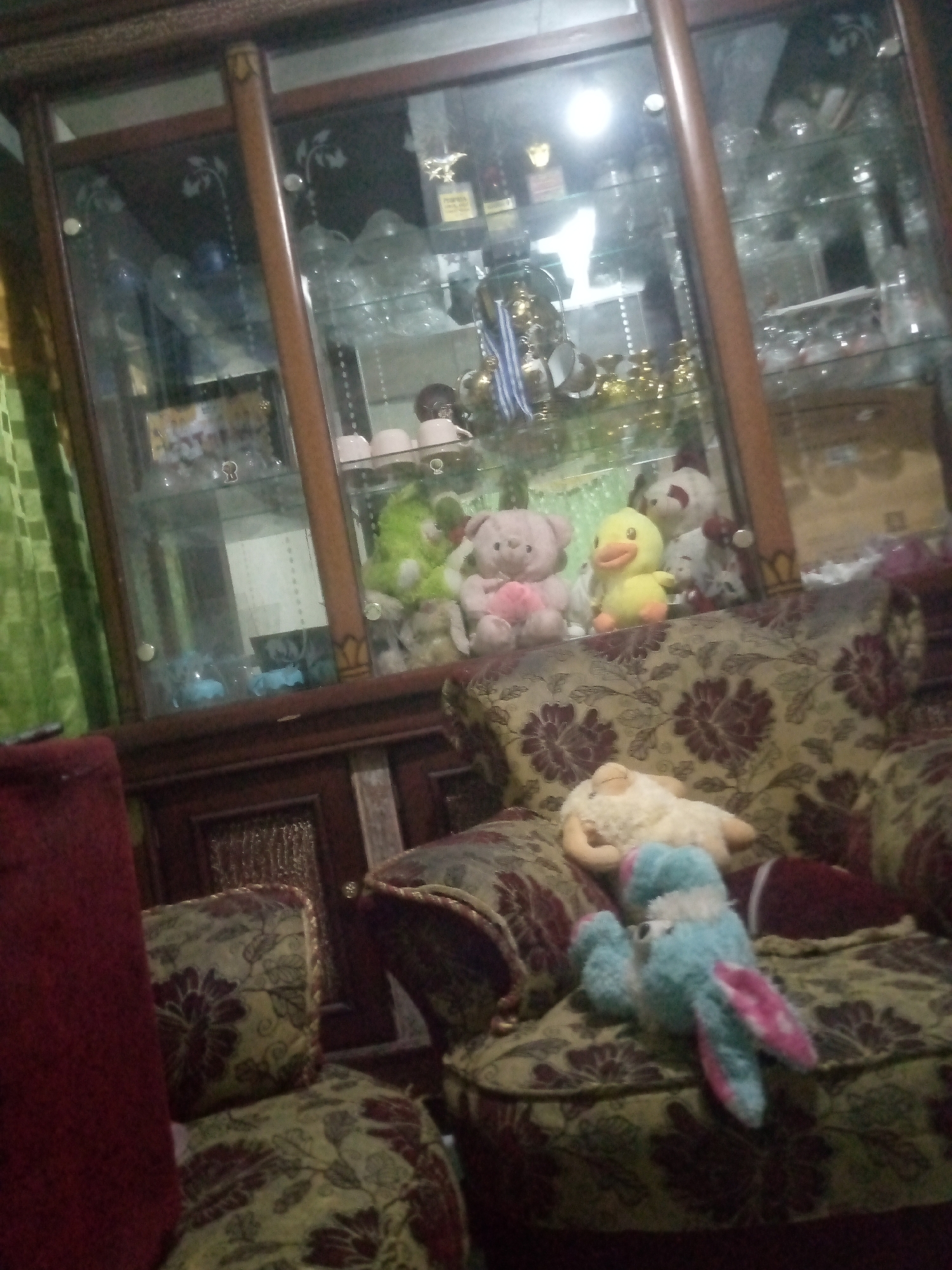 The image depicts an indoor scene, likely a living room, in Laksanamekar, Indonesia. The focal point is a large, wooden display cabinet filled with various items, including decorative cups, trophies, and what appear to be stuffed animals. To the right of the cabinet, a plush sofa with a floral pattern is visible, adorned with several stuffed toys. Among these are a pink teddy bear, a yellow duck, and a white lamb. On the left side of the frame, a portion of a red armchair is present, also covered in a patterned fabric. The lighting in the room appears dim, with a light source visible within the display cabinet.