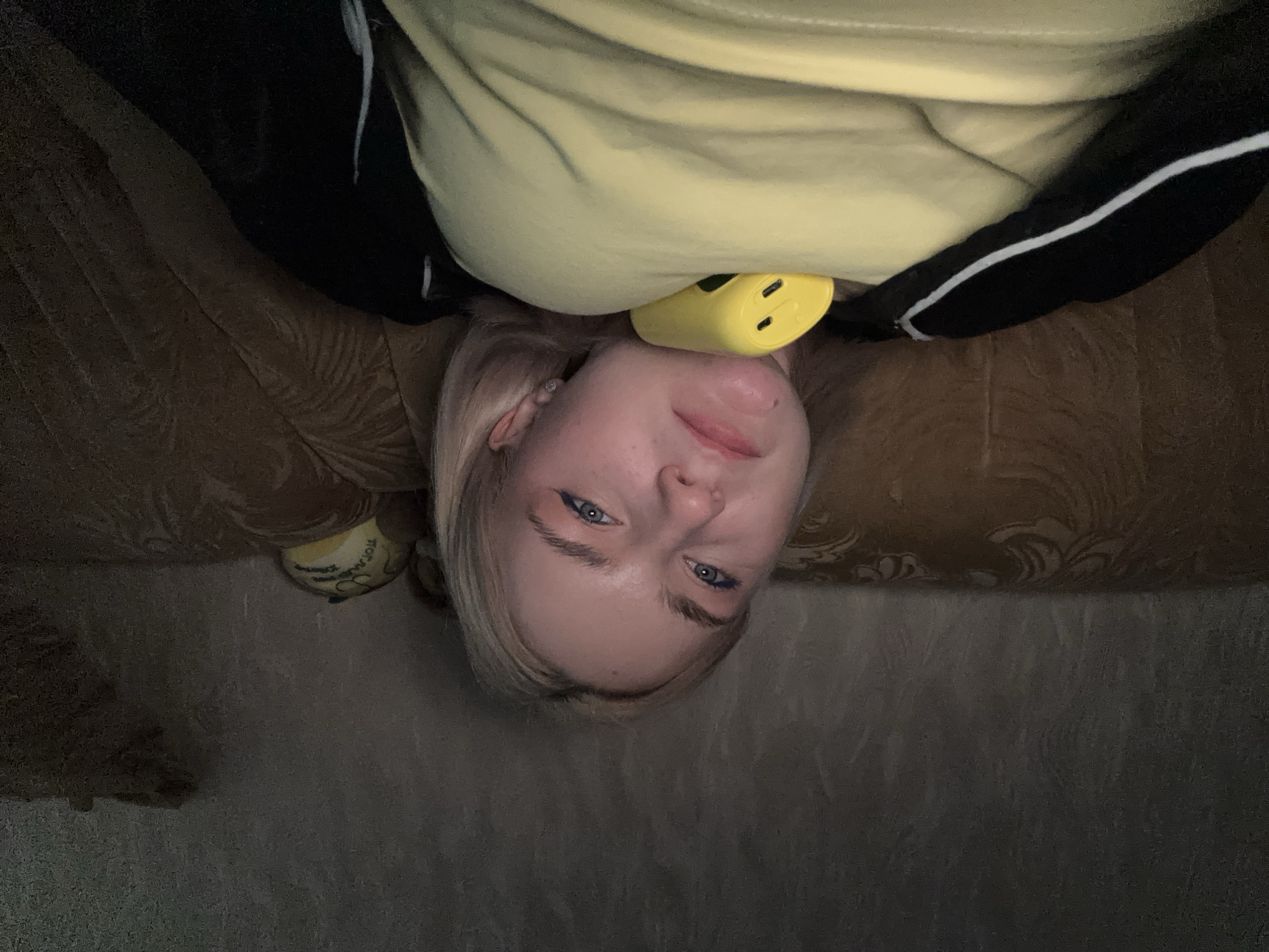 The image shows a young Caucasian woman lying down, with the camera positioned above her. Her head is at the bottom of the frame, and she is looking up at the camera with a slight smile. She has blonde hair, blue eyes, and is wearing a yellow t-shirt and a black jacket with white stripes. A small, yellow object, possibly a portable charger or a small toy, is visible tucked under her chin. The background appears to be a couch with a textured, patterned fabric. The lighting is dim, suggesting it might be indoors during the evening or in a poorly lit environment. There is no visible text in the image.