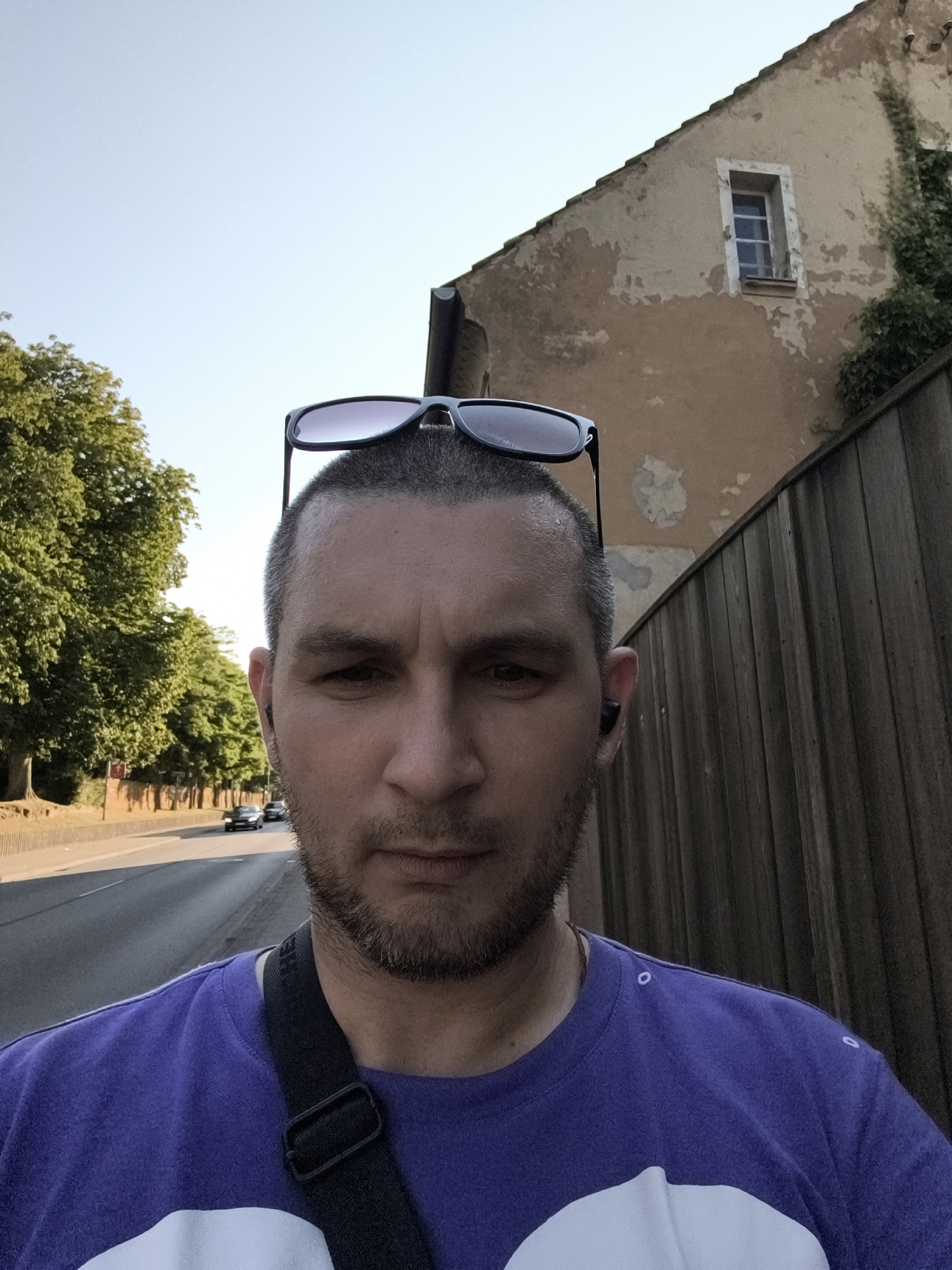 A man is photographed from the chest up, with his face centered. He has short, grey hair and a goatee. Sunglasses are perched on top of his head. He is wearing a purple t-shirt with a white graphic design. A black strap crosses his chest. He has a black wireless earbud in his right ear. The background shows a weathered building with a window, a wooden fence, and a tree-lined street with cars. The scene appears to be on the edge of town. The location is the city of Wolgast, Germany, at the intersection of Chausseestraße and Störtebekerstraße.