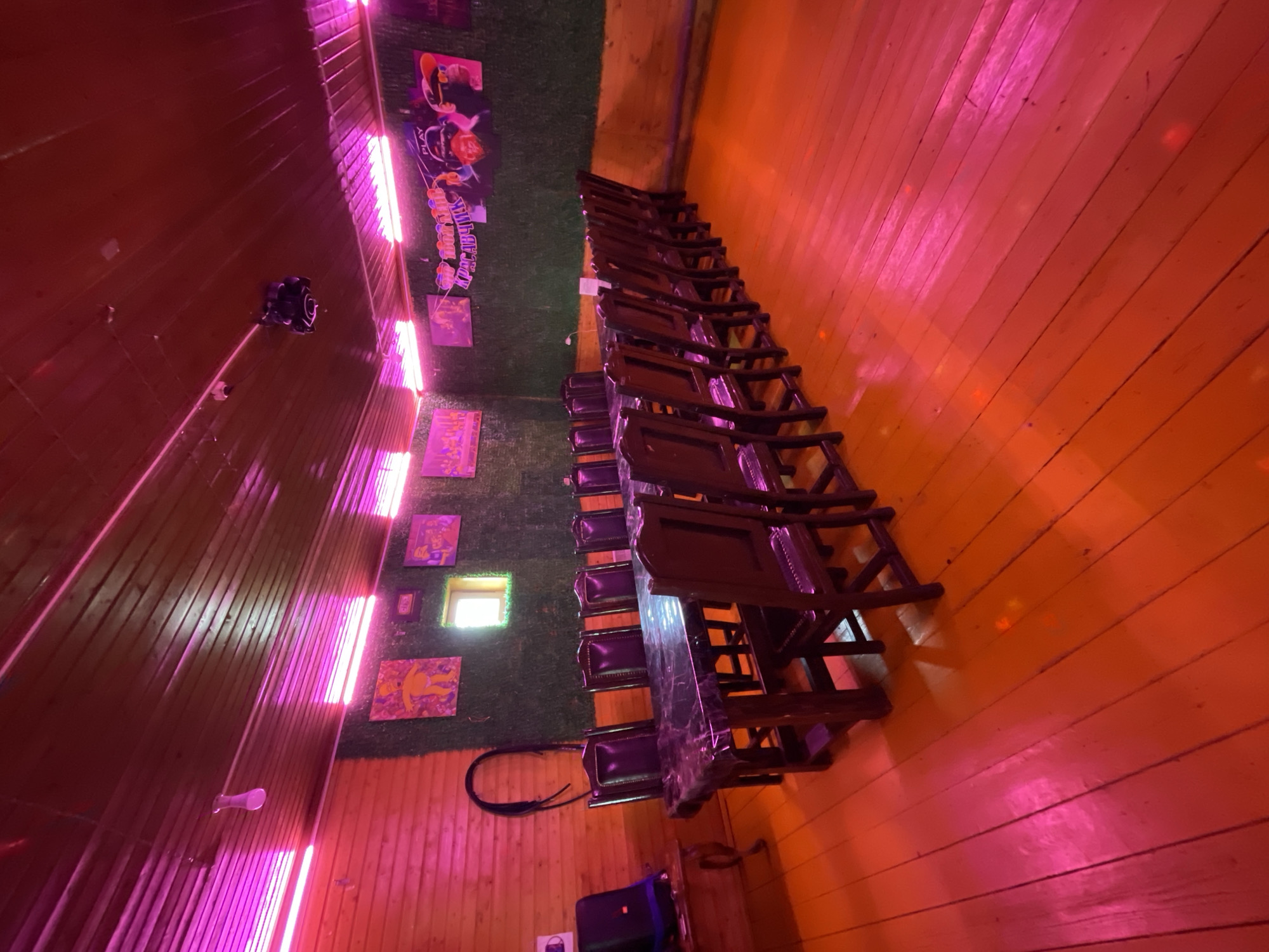 The image shows an interior scene in what appears to be a room with unique decor. Walls are wooden, illuminated with neon lights creating a pinkish-red hue. There is artificial turf on a portion of the wall adorned with framed pictures or posters. A line of stacked, dark-colored wooden chairs is placed near the transition between the wooden wall and the turf. A window is visible on the turf-covered part of the wall. The scene is likely indoors, but there are no people visible in the image. There is no indication of the city of Картмазово, Russia. The focus of the image is on the interior decor and the arrangement of the chairs.