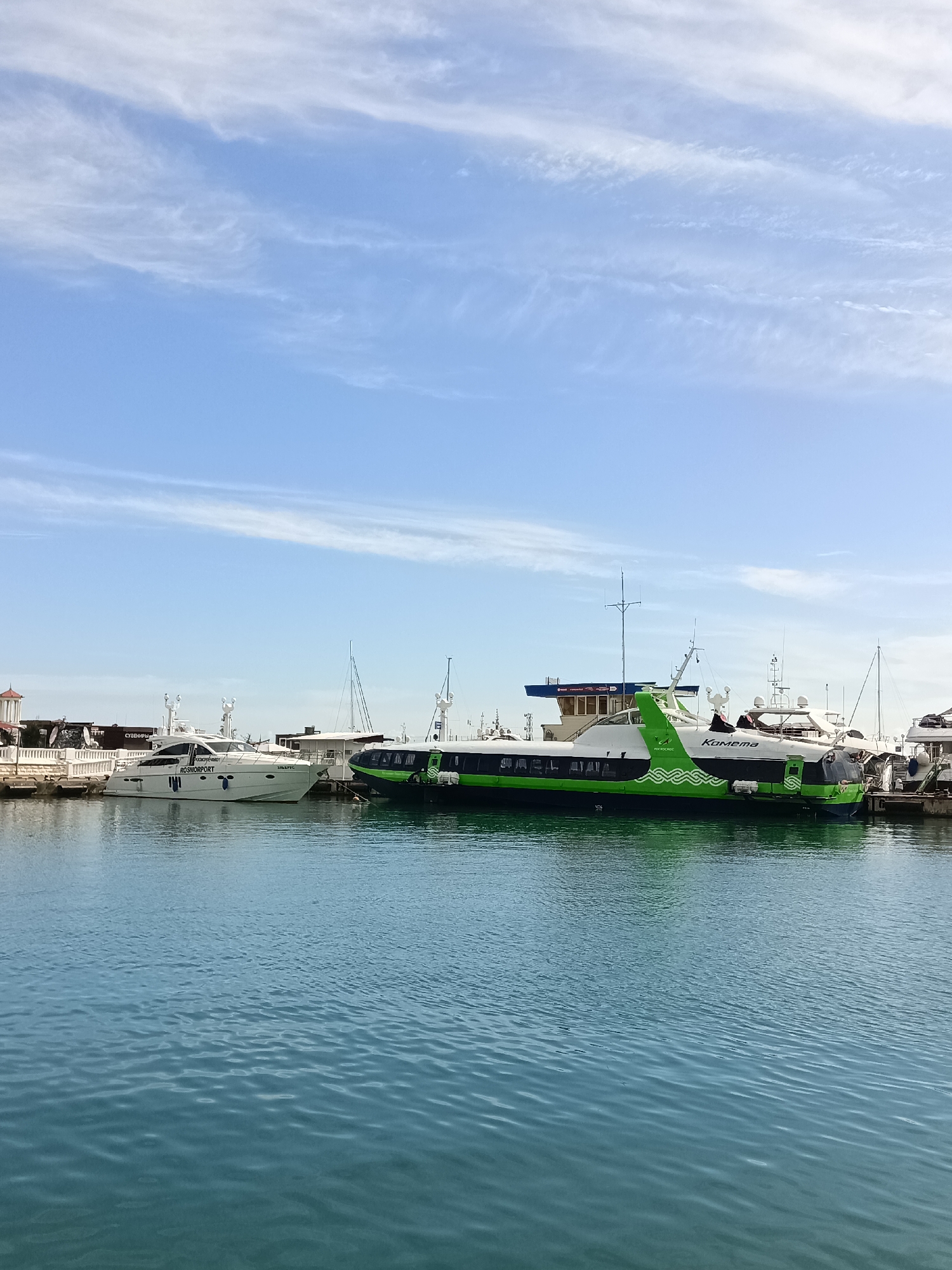 In Sochi, Russia, a bright, partly cloudy sky is visible above a body of water reflecting the sky. Moored in the foreground are two vessels: a white yacht with the word "ROSMORPORT" emblazoned on its side, and a green and white hydrofoil with the word "Комета" (Kometa) written on its side. The hydrofoil is larger than the yacht and appears to be in operational condition. The waterfront is lined with structures, including what appears to be a building with a red-tiled roof and white railings, and various masts of other boats can be seen in the background. The water is a deep blue-green and exhibits a slight ripple.