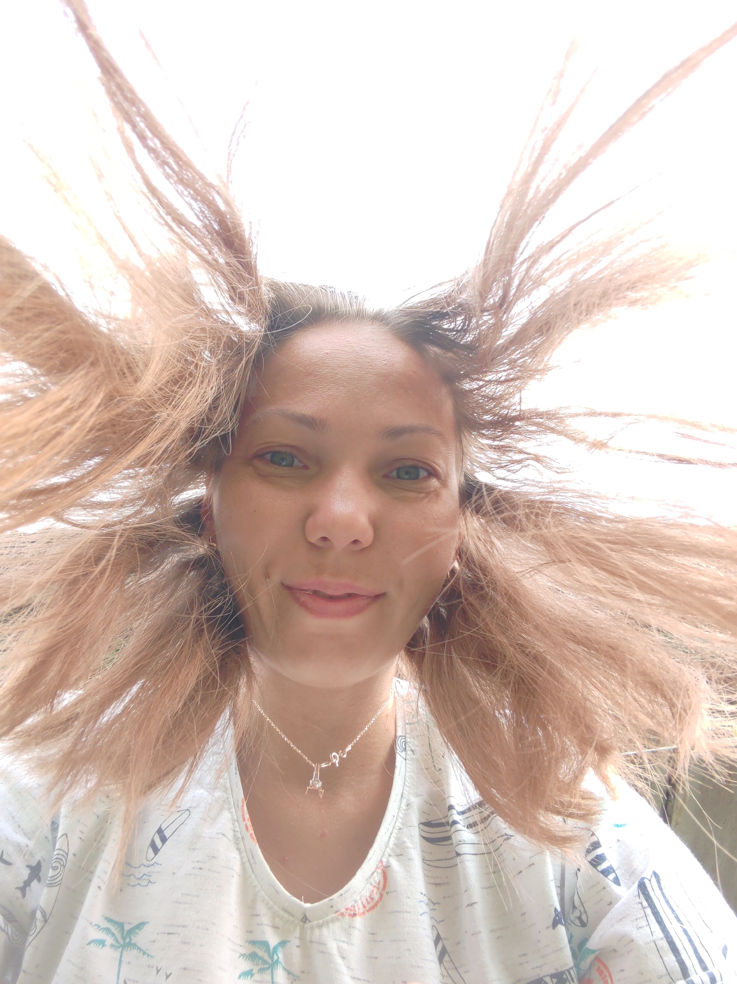 A woman's face is captured from a low-angle perspective, with her blonde hair dramatically fanned out around her head by an unseen force, likely wind. She appears to be outdoors, with a bright, washed-out background that suggests daylight. Her eyes are open and looking towards the camera, and she has a slight smile. She is wearing a light-colored V-neck shirt with a pattern of palm trees and surfboards. A delicate silver necklace with a small charm is visible on her collarbone. The image is underexposed, especially in the upper portion, due to the bright light source. There is no discernible location in the city of Чухлэм, Russia, visible in the image.