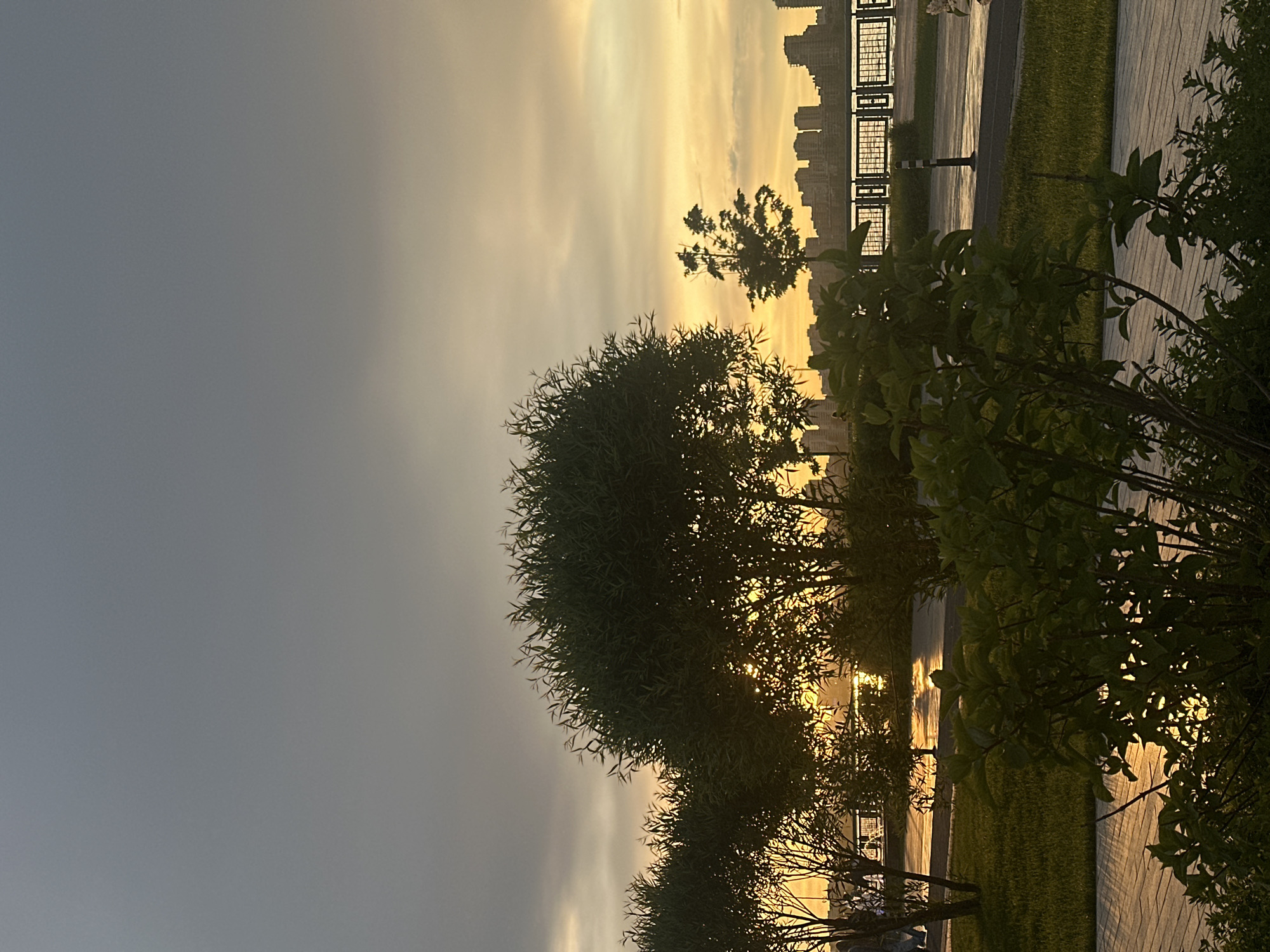 The image shows a view of the city of Kazan, Russia, at sunset. The sky is a mix of gray and yellow, with the sun setting behind the buildings in the distance. In the foreground, there are trees and bushes, and a sidewalk runs along the edge of a grassy area. There are no people visible in the image.