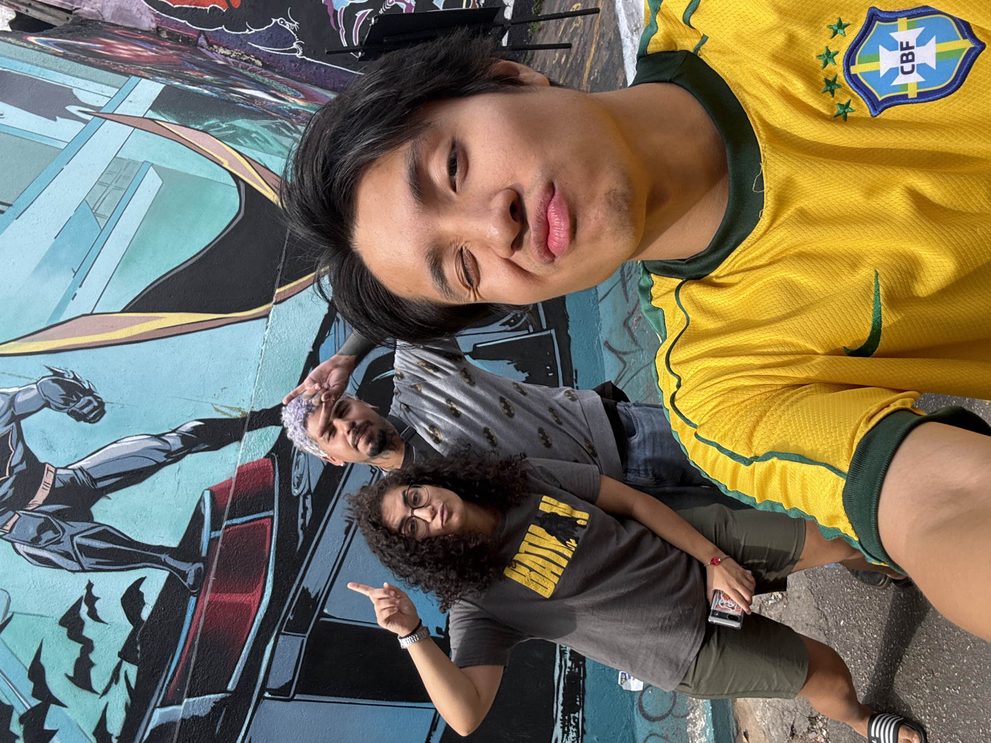 Three people are posing for a selfie in front of a mural. The person taking the selfie is wearing a yellow and green soccer jersey with a Brazilian emblem. The person in the middle has lavender hair, a beard, and is wearing a grey t-shirt with a batman pattern. The third person is wearing glasses and a grey t-shirt with a yellow bat symbol and is pointing at the Batman mural. The mural is a depiction of Batman jumping out of a Batmobile. The scene is on a city street in São Paulo, Brazil.