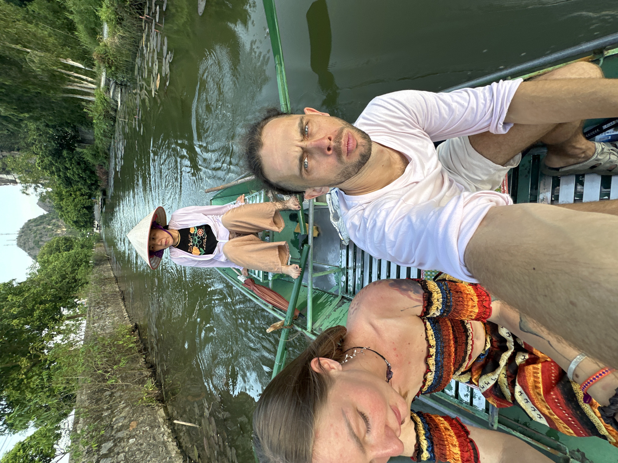 Three individuals are seated in a green boat navigating a narrow, murky waterway lined with vegetation and a stone wall. The person in the foreground, a man with blue eyes and a beard, is looking directly at the camera, his mouth slightly pursed. He is wearing a light pink long-sleeved shirt and white shorts. To his left, a woman with brown hair and fair skin is looking down, her head tilted. She is wearing a colorful off-the-shoulder top with ruffled sleeves and a patterned skirt. Her hair is partially covering her face. In the background, a third person, wearing a conical hat, a black t-shirt with a colorful floral design, and tan pants, is seated further back in the boat, also looking forward. The boat is propelled by a wooden oar. The waterway appears to be in a rural or natural setting, with trees and hills visible in the distance. The overall lighting suggests it is either early morning or late afternoon. This scene is consistent with boat tours in the karst landscape of Ninh Binh province, potentially near Hoa Lư, Vietnam, known for its rivers and limestone formations.
