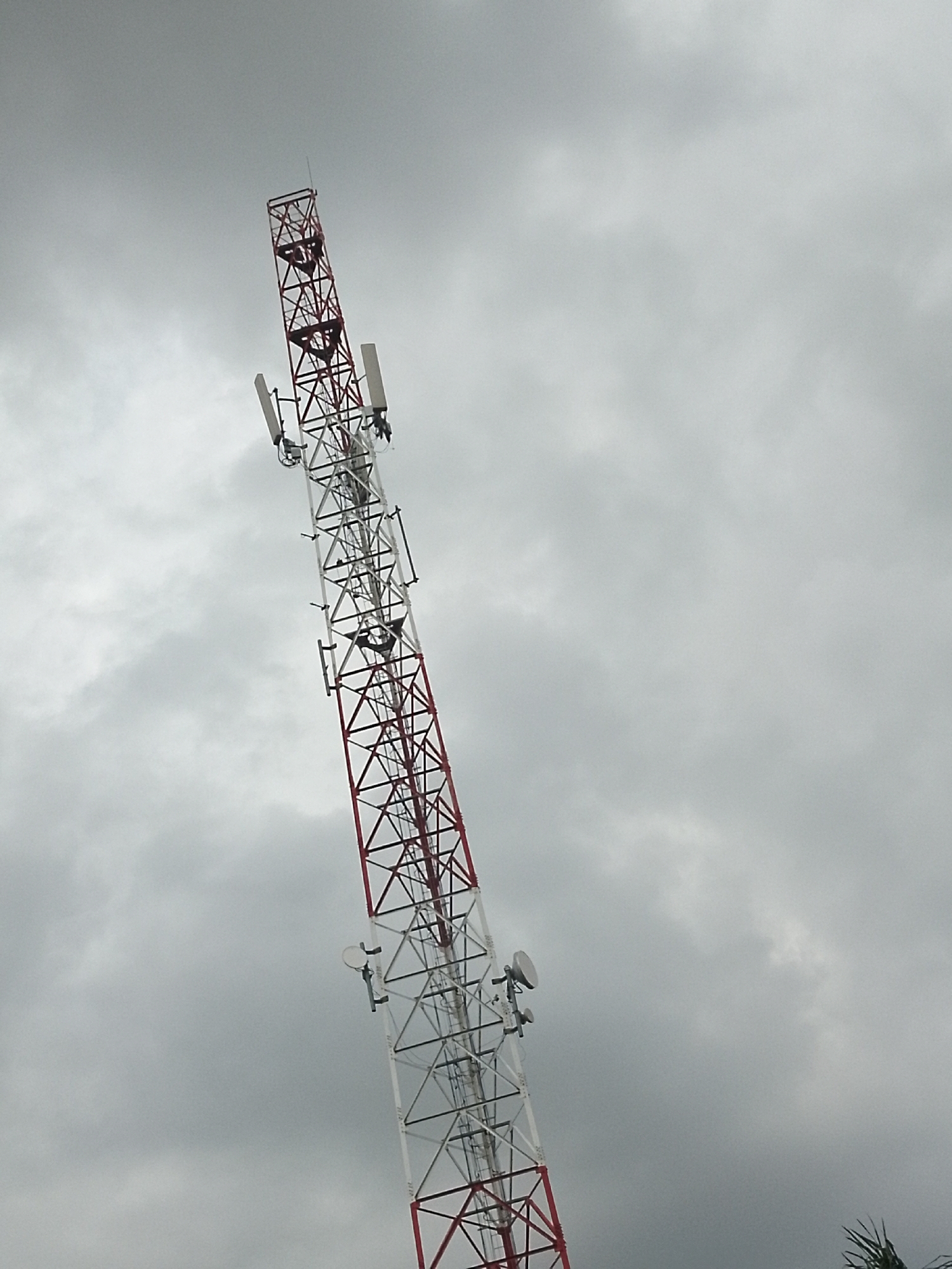 An outdoor, low-angle, full shot captures a cell tower against a cloudy sky. The tower, a lattice structure painted red and white, dominates the right side of the frame, extending vertically upwards. Attached to the tower are various antennas and dishes. The sky is overcast with grey clouds. The image provides no identifiable landmarks or context to ascertain its specific location within Serti, Nigeria, or any other city.