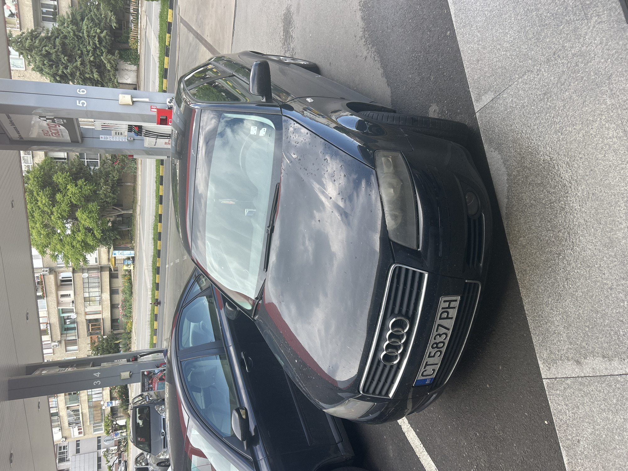 A black Audi A4 with license plate "CT 5837 PH" is parked at a gas station in Kazanlak, Bulgaria. The car is positioned in front of gas pumps numbered 5 and 6. Another car is parked closely behind it. The scene includes buildings across the street from the gas station and surrounding vegetation.