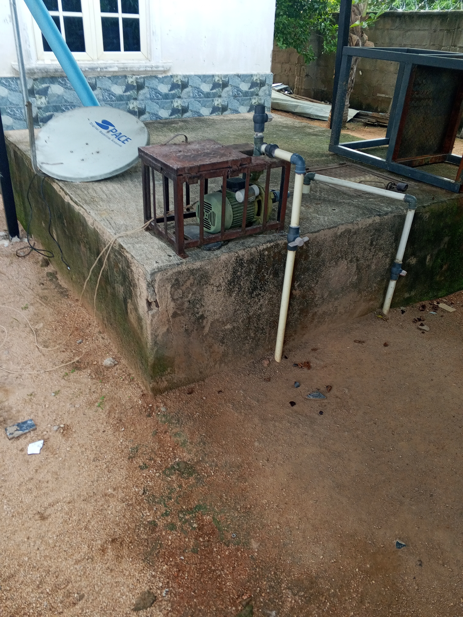 In the city of Howayi, Nigeria, a satellite dish branded "SPACE" is positioned on a concrete platform next to a green motor enclosed in a metal cage. Light blue piping connects to the motor, extending upwards and then horizontally. The platform itself is aged and weathered, with patches of moss. Surrounding the platform is sandy, uneven ground with scattered debris and small stones. In the background, a building with white walls and blue tiled accents can be seen, along with a large metal structure. The overall scene depicts an outdoor utility area with visible signs of use and exposure to the elements.