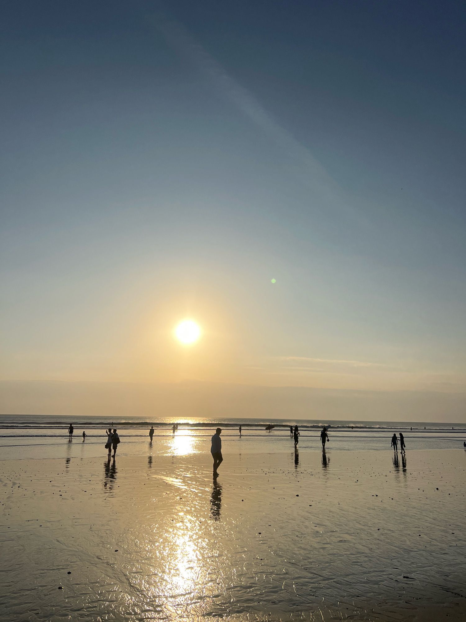 The image depicts a sunset scene on Kuta Beach, Bali, Indonesia. Silhouetted figures of people are visible walking and standing on the wet sand. The sun is low on the horizon, casting a bright, golden reflection on the water and wet sand. The sky is clear with a subtle gradient from a light blue at the top to a warmer yellow near the horizon. Small waves are breaking in the distance. The wet sand shows intricate patterns from the receding tide.