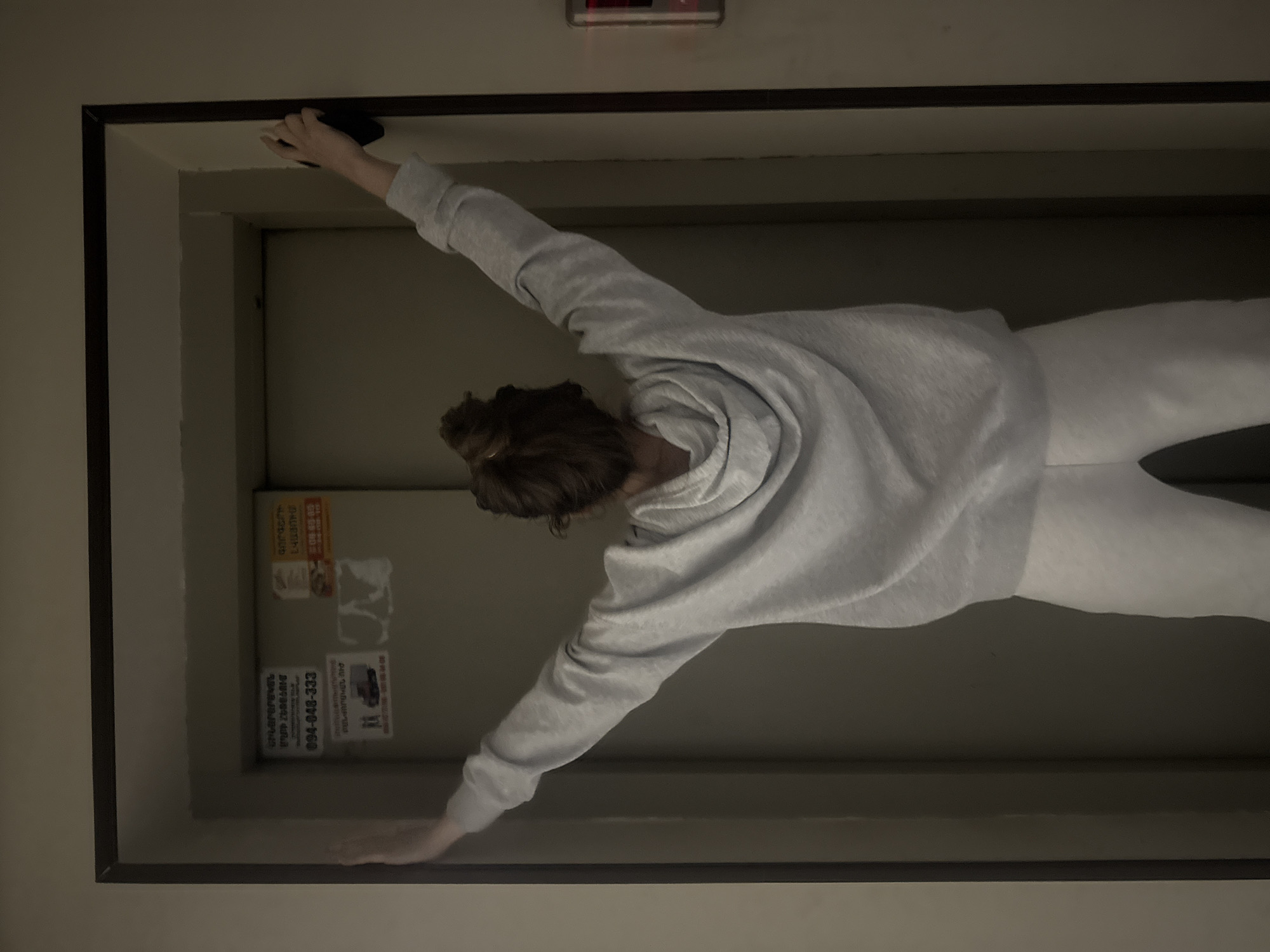 A person wearing a gray hoodie and white pants is standing in what appears to be an elevator. They are positioned in the center of the frame with their arms outstretched, touching the top and bottom of the elevator door frame. The elevator interior has beige walls, and the back wall contains stickers and advertisements. A red rectangular object is visible at the top of the elevator. The location of Yerevan, Armenia is inferred from the presence of the number "094" on a sticker that is visible on the back wall.