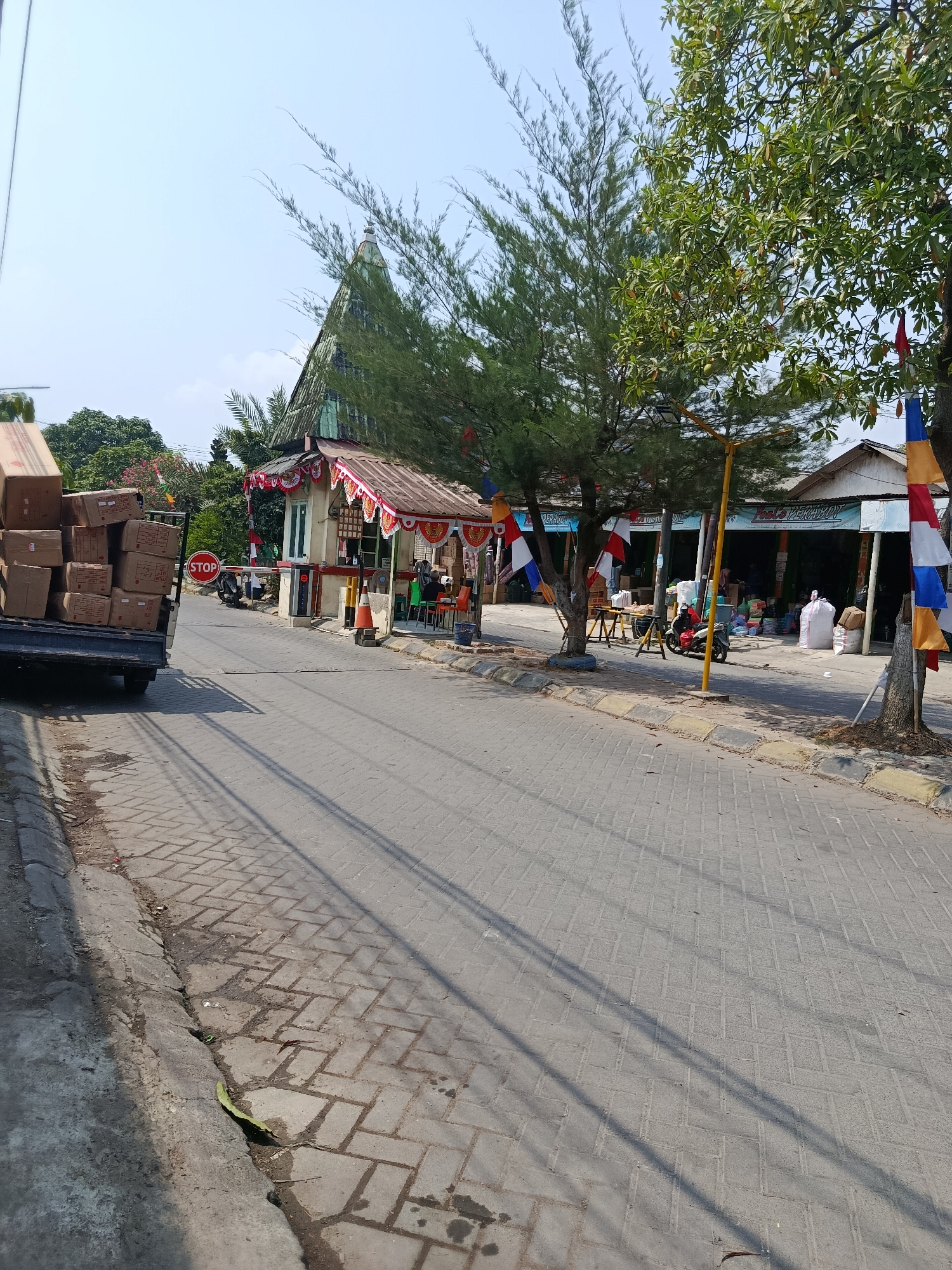 The image captures a street scene in Wanasari, Indonesia, likely a commercial area. In the foreground to the left, a pickup truck is loaded with cardboard boxes. A red stop sign is visible on a pole in the middle ground, next to a small building that appears to be a guard post or toll booth. This building is decorated with red and white festive streamers.
Beyond the guard post, a row of small shops lines the street. One shop has a sign that reads "TOKO PERAMY". The shops display various goods, including sacks of what appear to be rice or other staples, plastic containers, and other merchandise. A person on a red and black motorcycle is seen riding past the shops.
Several trees are present, with a tall evergreen tree with needle-like leaves dominating the center of the frame. Other trees with broader green leaves are also visible. Indonesian flags, in red and white, are hung along the street, indicating a festive or national occasion. The road is paved with interlocking bricks, and the sky is clear and blue, suggesting a sunny day. The overall impression is of a bustling, somewhat informal marketplace or roadside commercial strip.